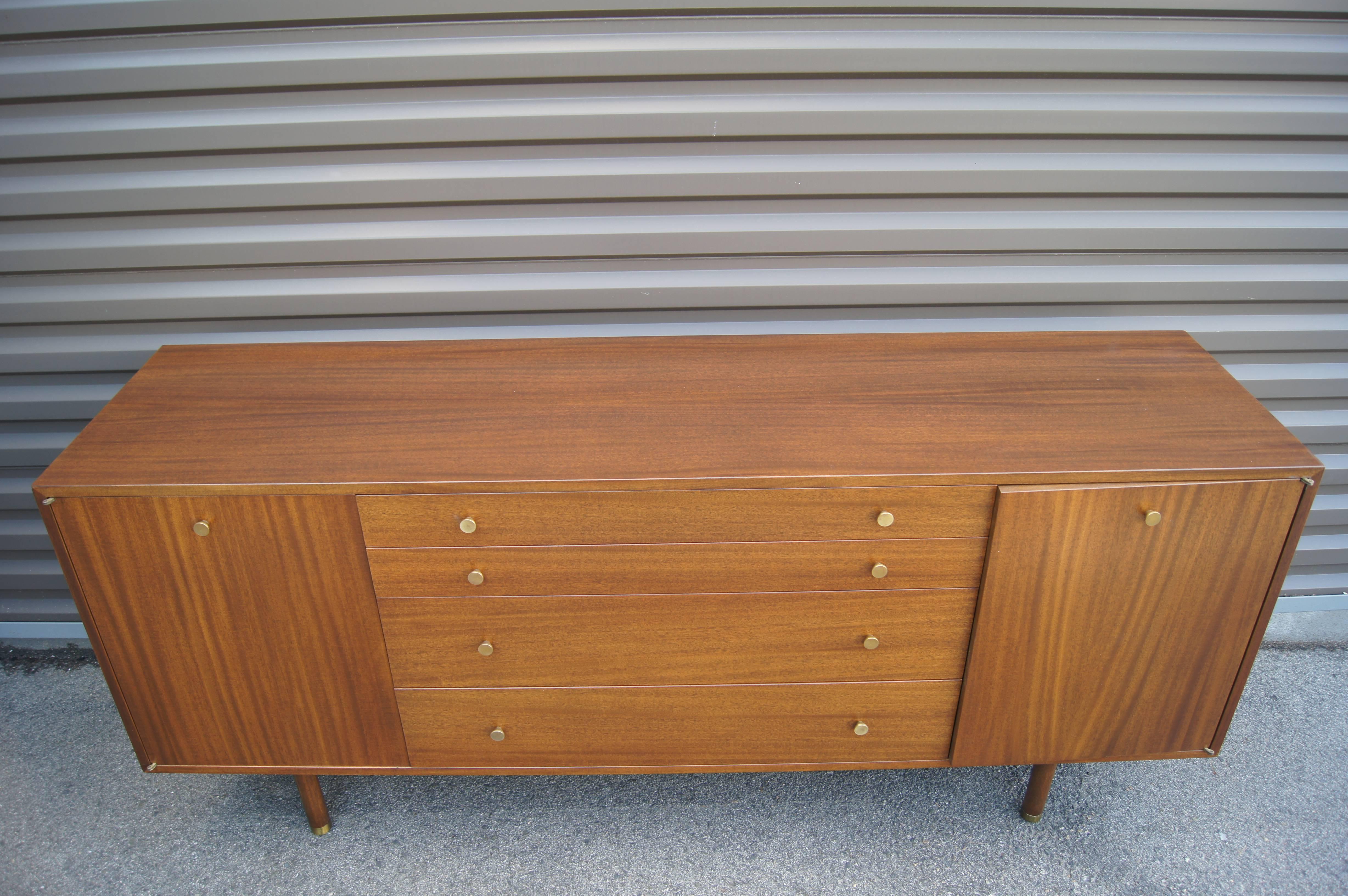 Designed by Harvey Probber, this midcentury credenza features a gorgeous mahogany grain complemented by brass pulls and brass-capped legs. Doors on either end conceal adjustable shelves. In the center are two shallow drawers, one felt-lined and