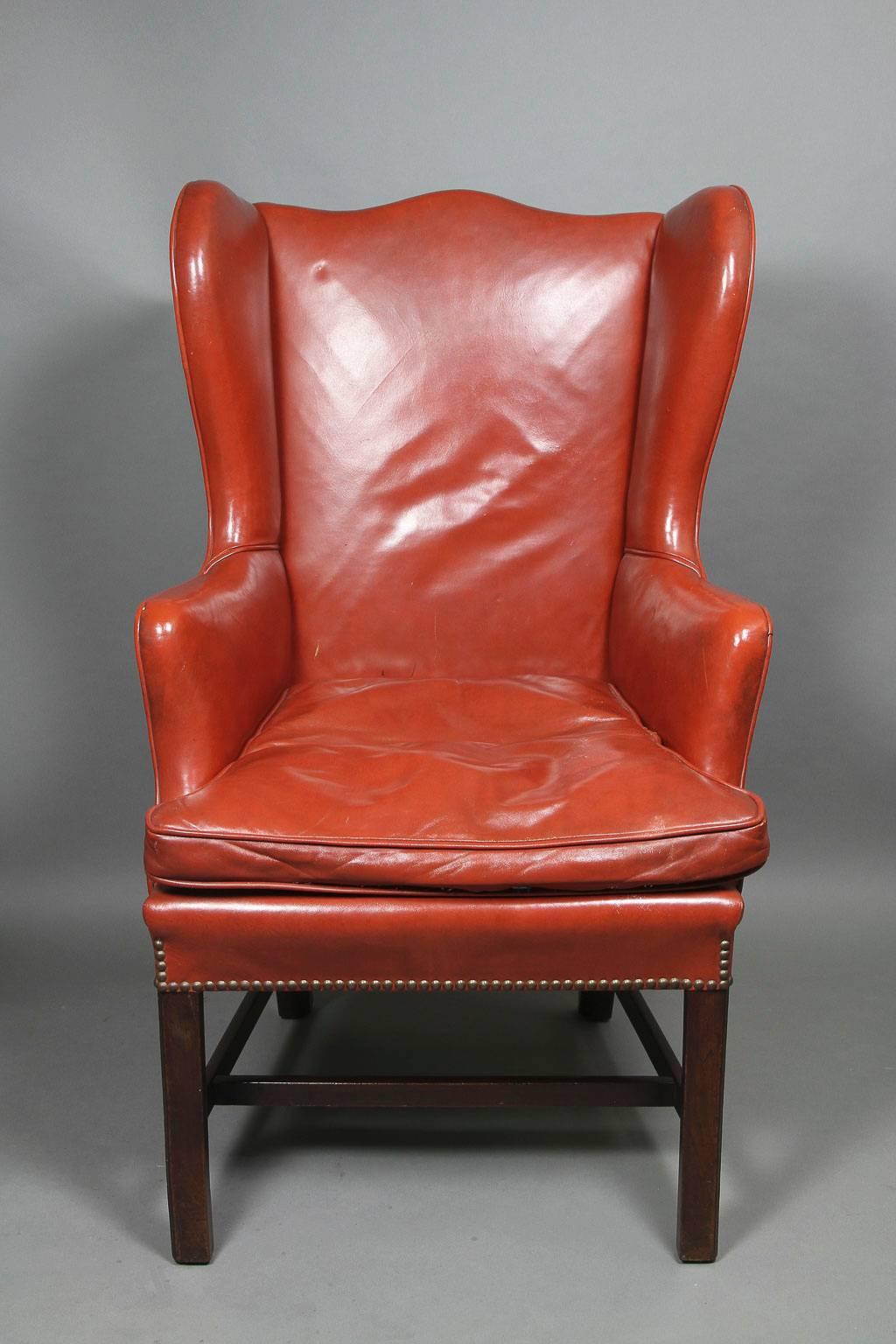 Simple form with serpentine crest and shaped wings, scroll out arms and loose cushion seat, upholstered in leather with brass tacks, raised on square section legs and H form stretcher.