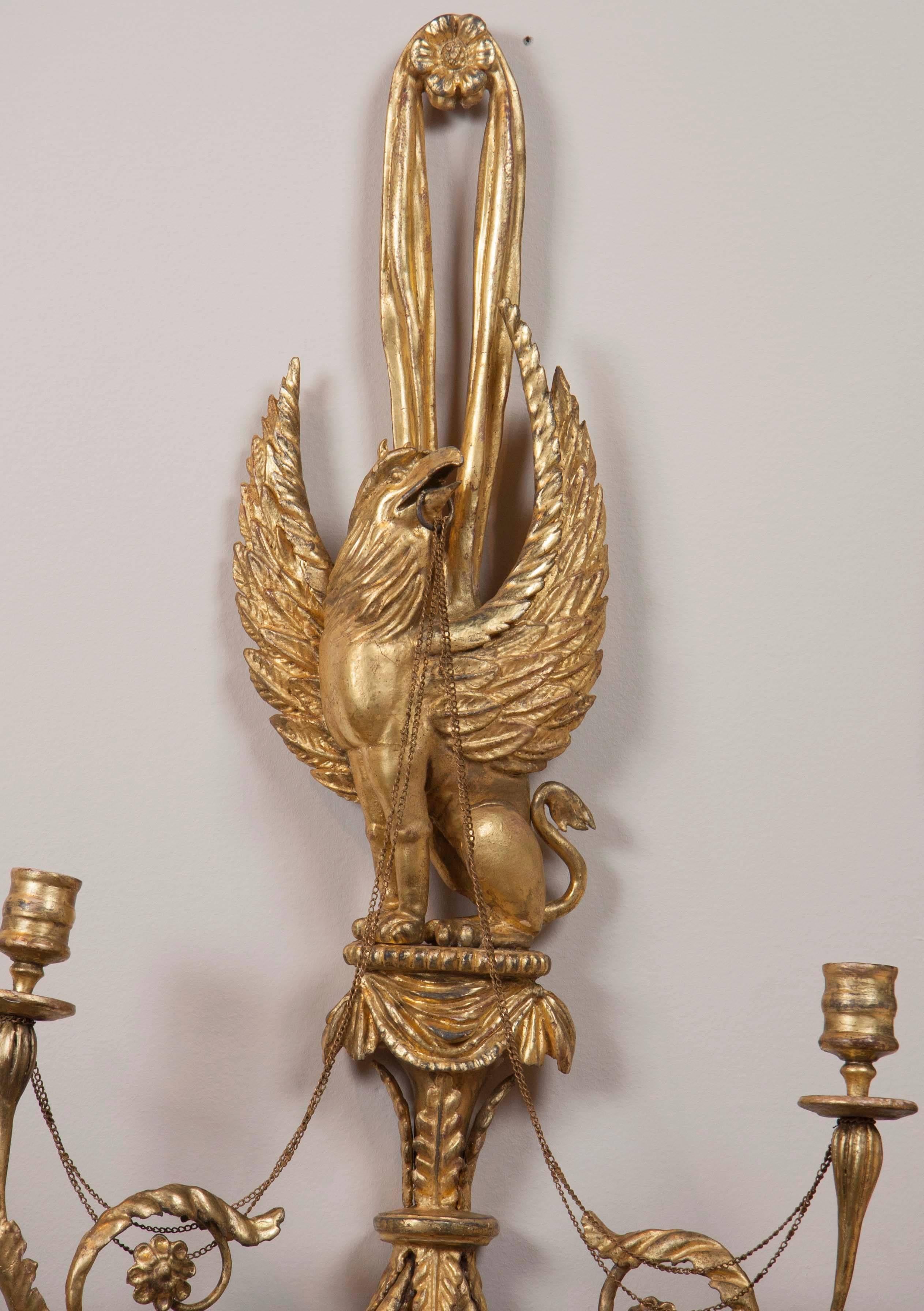 Each features a gryphon with a ring in its mouth and chains connecting to two taper candle holders, with details of leaves and flowers. Has been re-gilded. Carved wood.