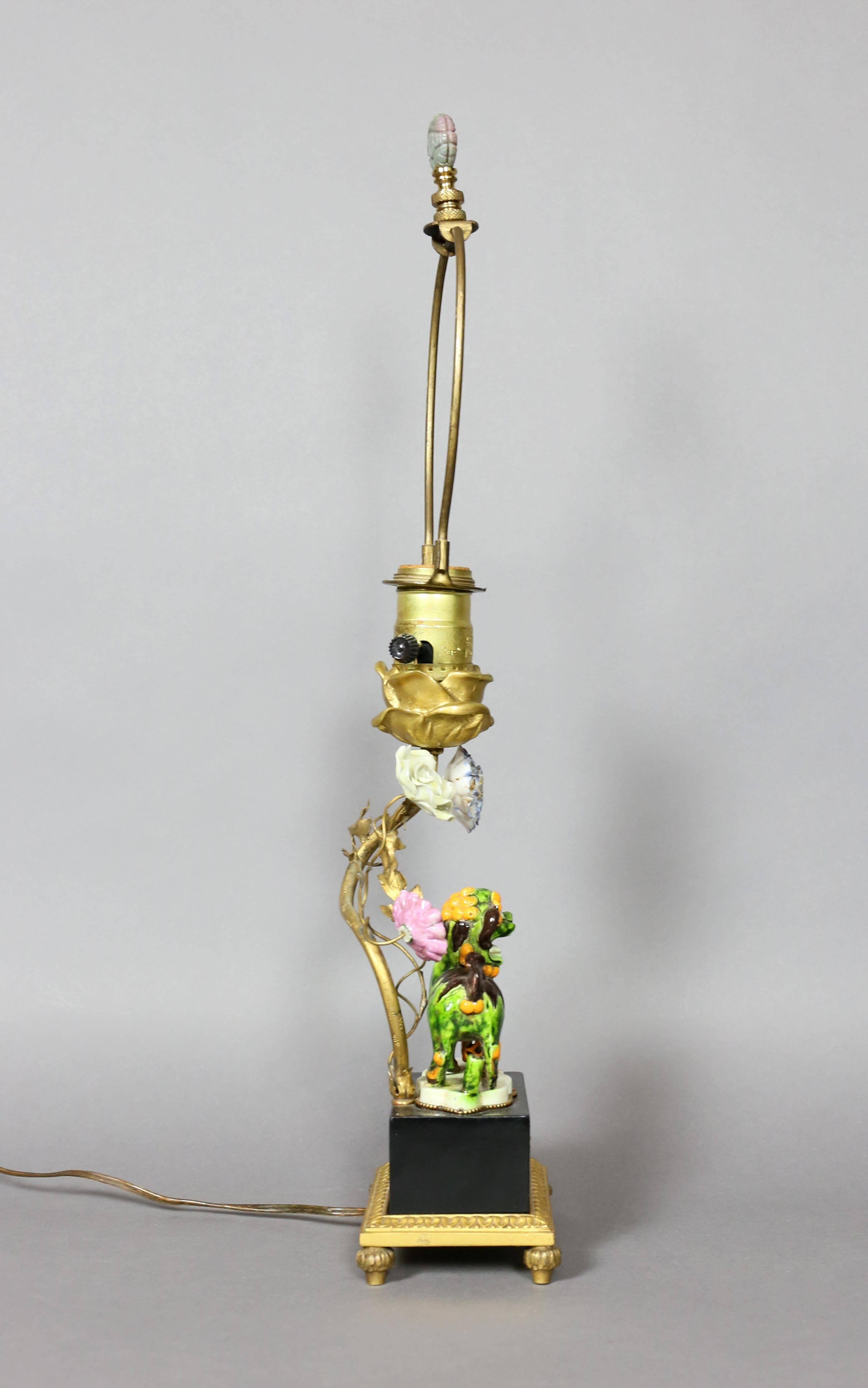The pooch painted in greens, browns and orange with paw on a pierces ball, cartouche shaped base, mounted on an ebonized base with enamelled cartouche, leaf tip bronze feet. Light stem with mounted porcelain flowers.