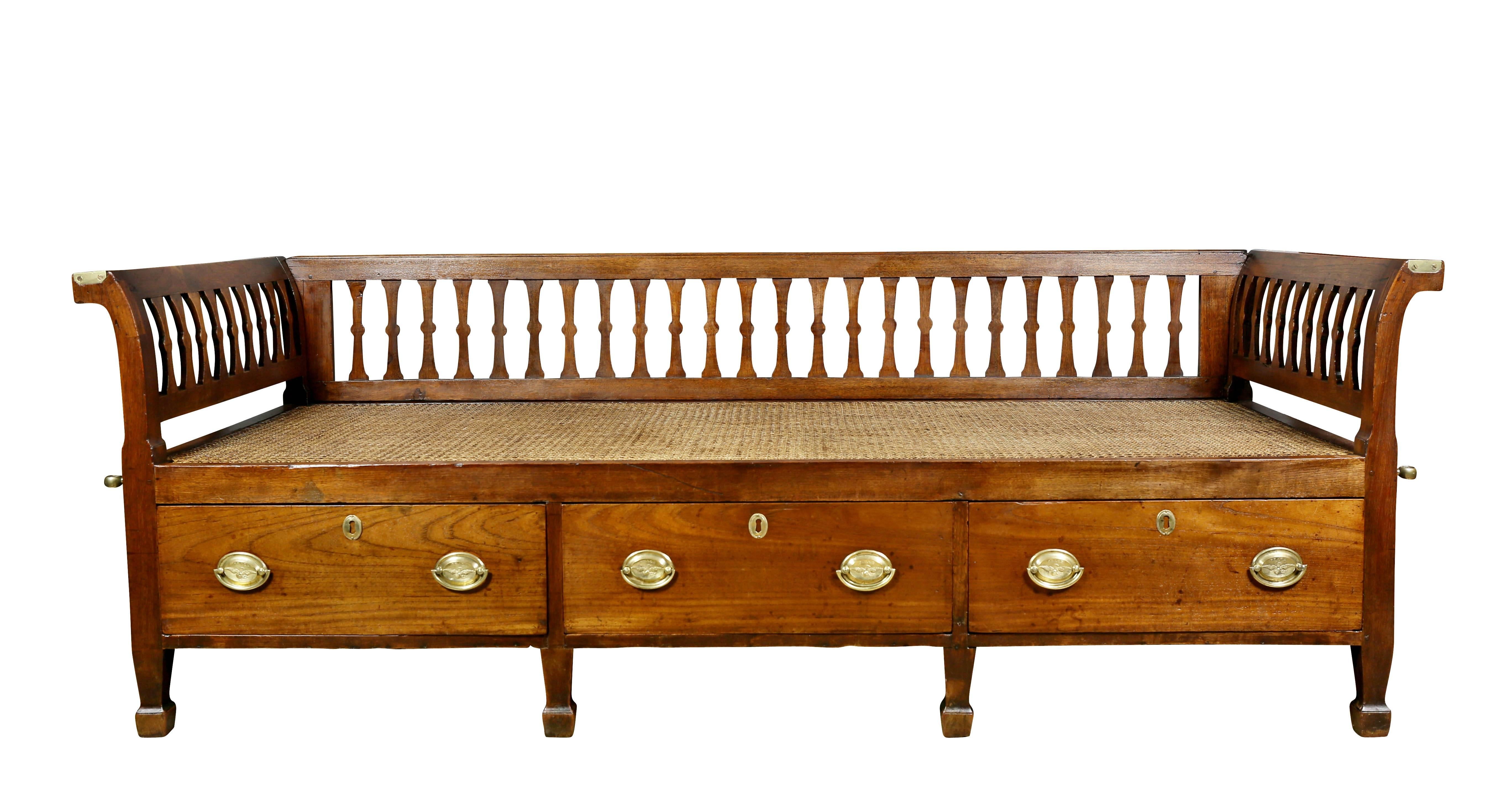 With detachable slat back and slightly scrolled arms over a caned seat over three drawers, raised on square tapered legs. Carrying handles on sides and brass fittings presumably for mosquito netting or canopy.