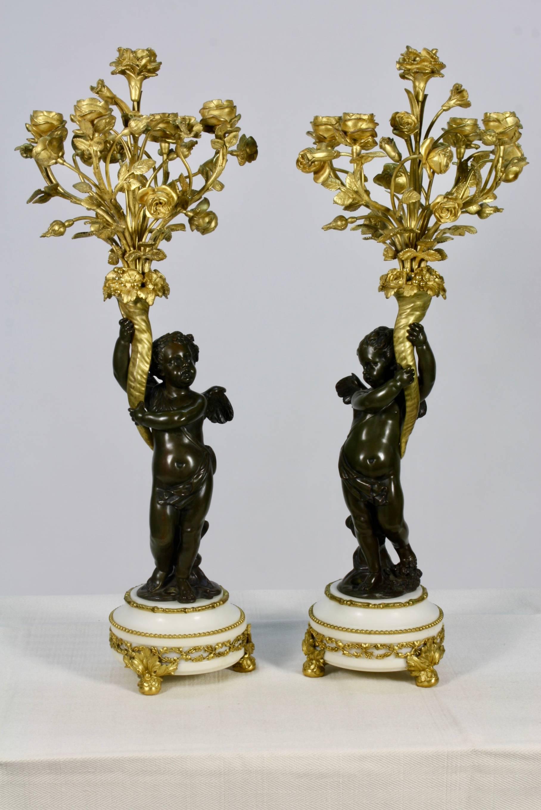 Pair of candelabra with winged putti, each supporting a floral bouquet, on top of marble base. Each putti is patinated bronze and the bouquets are gilt bronze. The floral bouquet has five candle cups. The candle cups and bases have been drilled for