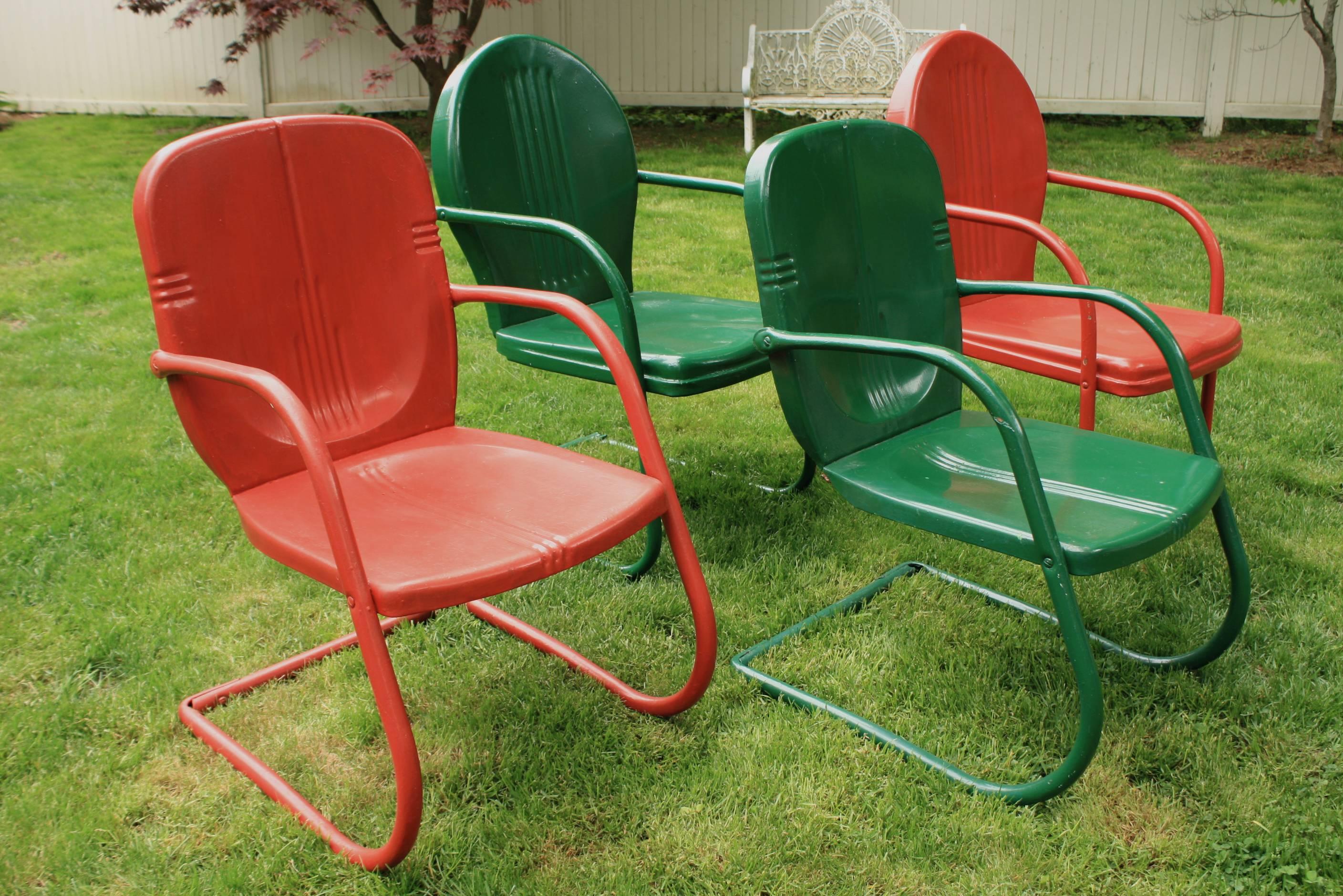 A set of four painted metal garden or patio armchairs, consisting of two pairs: one pair taller with rounded or "lollipop" backs, and the other pair shorter with flatter more deco-styled backs). Tubular arms and legs create a comfortable