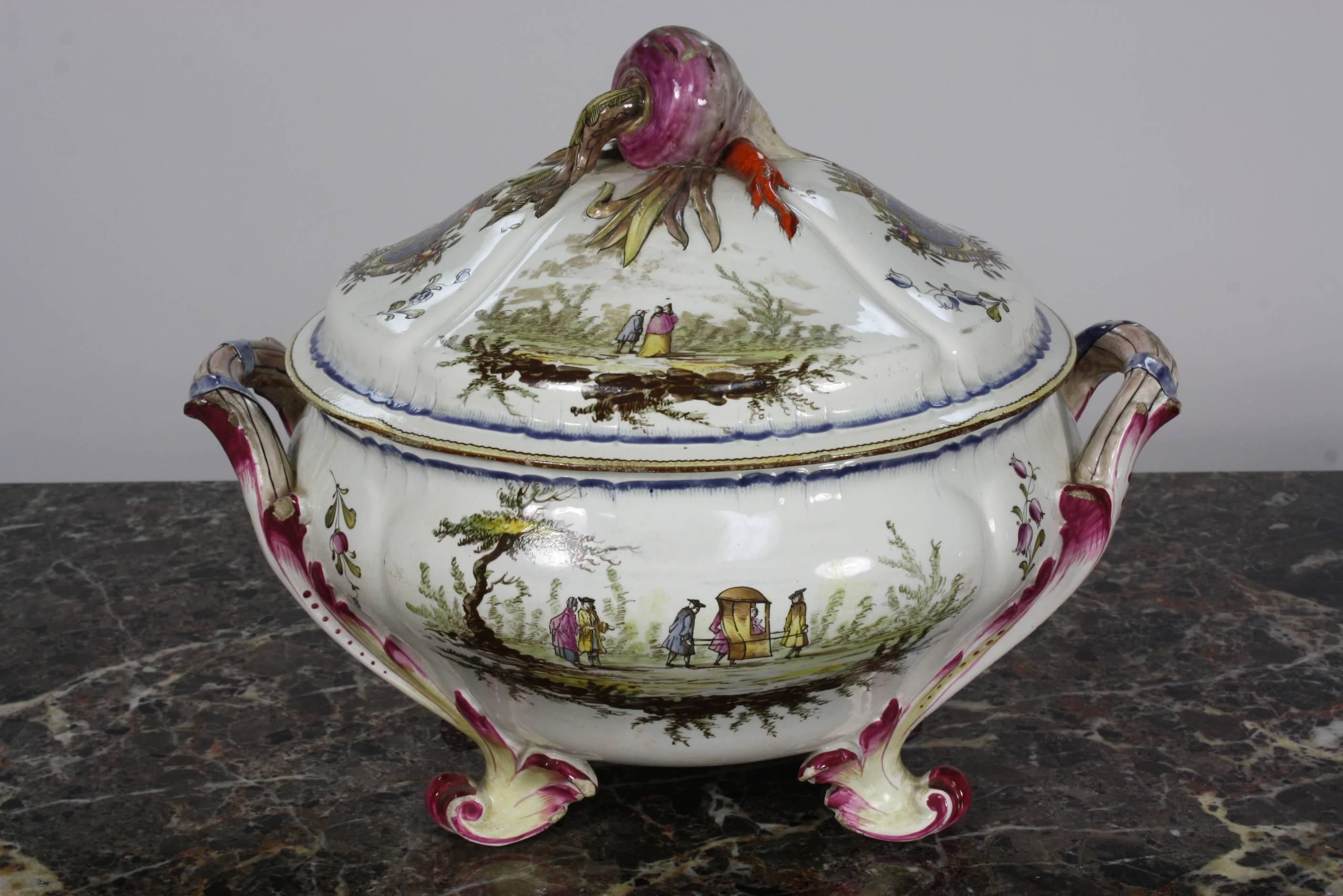 A lovely, highly-detailed French faience soup tureen, with handles, feet and nicely-sculpted vegetable decorated cover (18th century). The top cover has a radish, carrot and scallion. The painted decoration on the sides includes a shield with