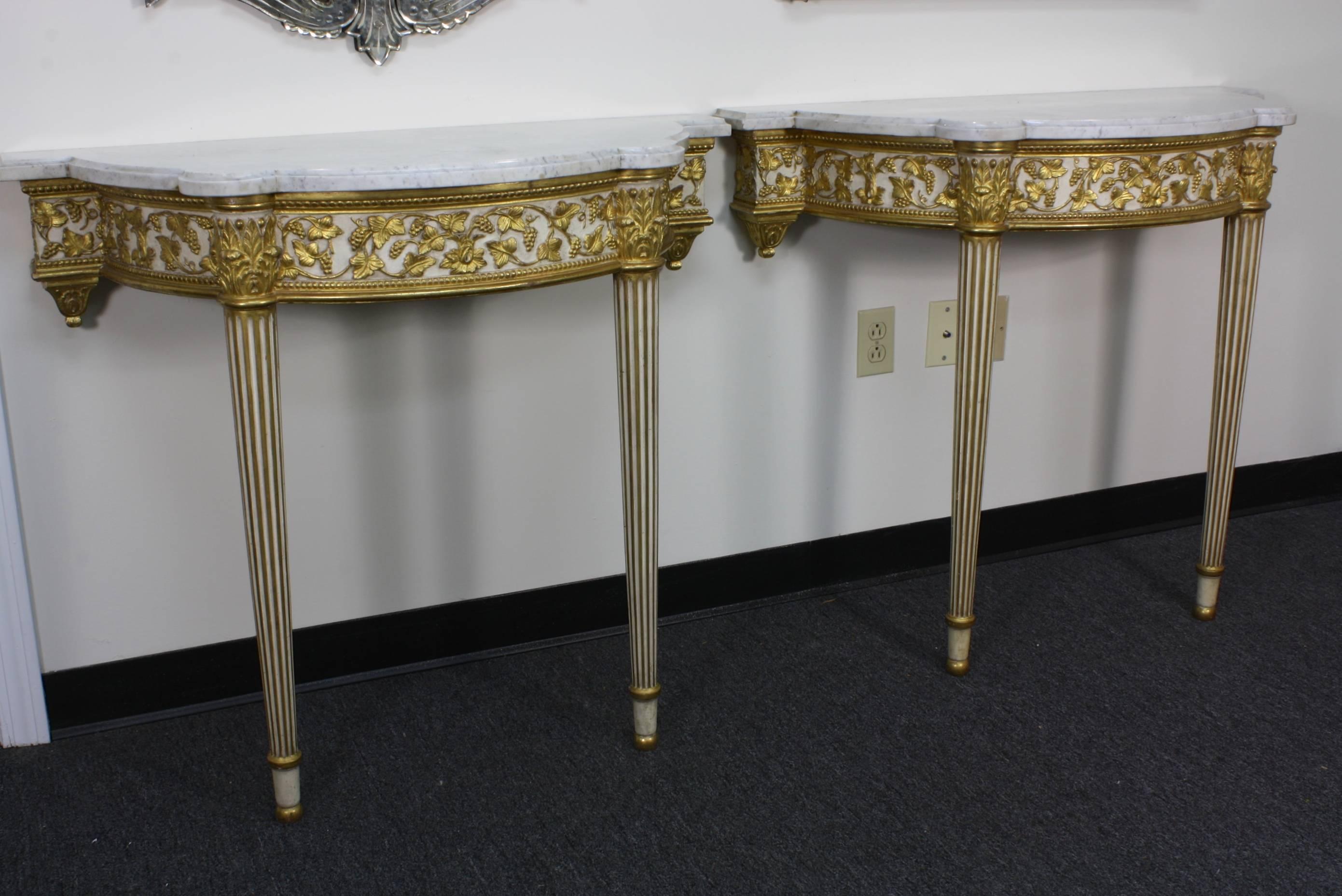 A lovely pair of French white-painted and gilded console tables, with Carrara marble tops, in the Louis XVI style (early 20th century). The raised gilded decoration is highly-detailed, featuring grape vines and leaves, and the legs are tapered and