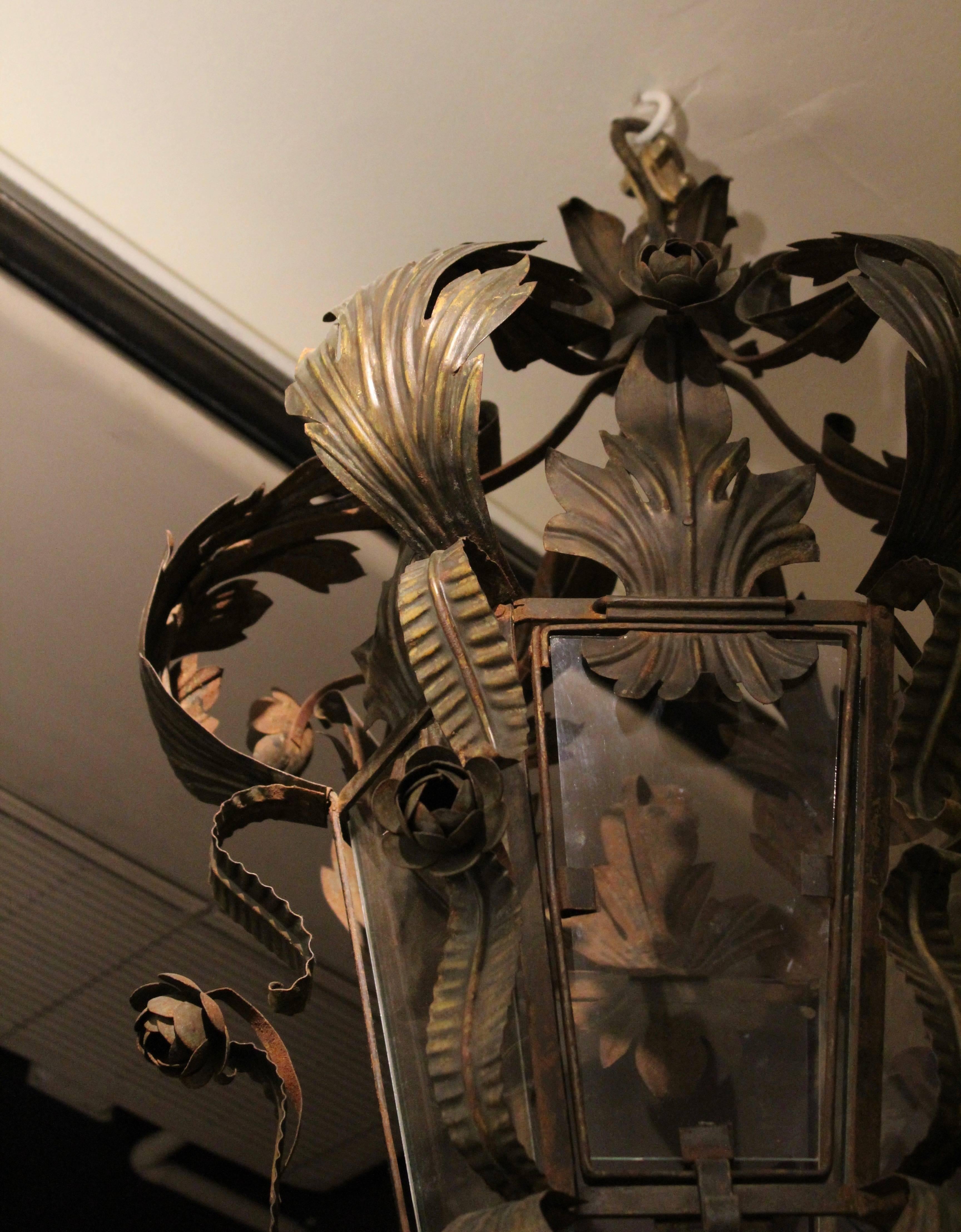 French Provincial wrought iron lantern in a foliate design, circa 1930. The top of the lantern formed by large curving acanthus leaves interspersed with roses, above the hexagonal glass body, each edge with a scrolling leaf centred by a rose. The