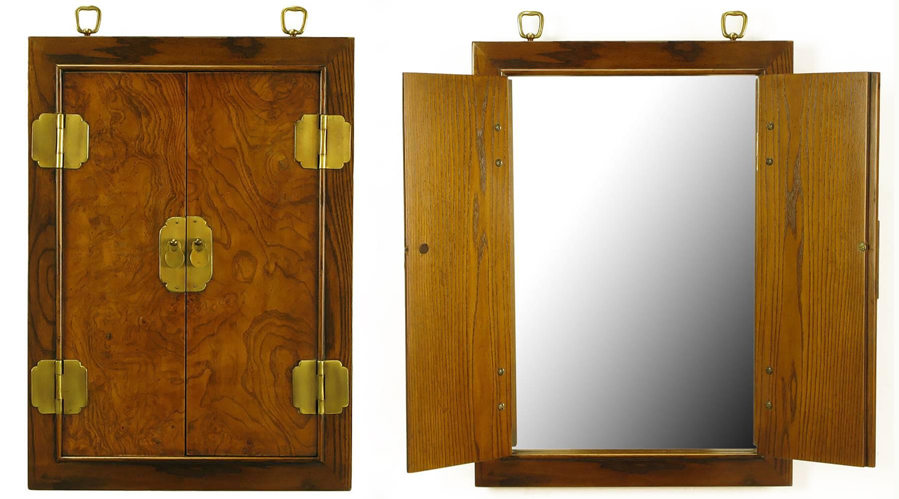 Uncommon two-door enclosed handing wall mirror with exposed brass hinges, brass escutcheons and Asian Style drop disc brass pulls. The doors are covered in burled walnut and the piece s framed in ashwood. Topped with brass ring hangers.