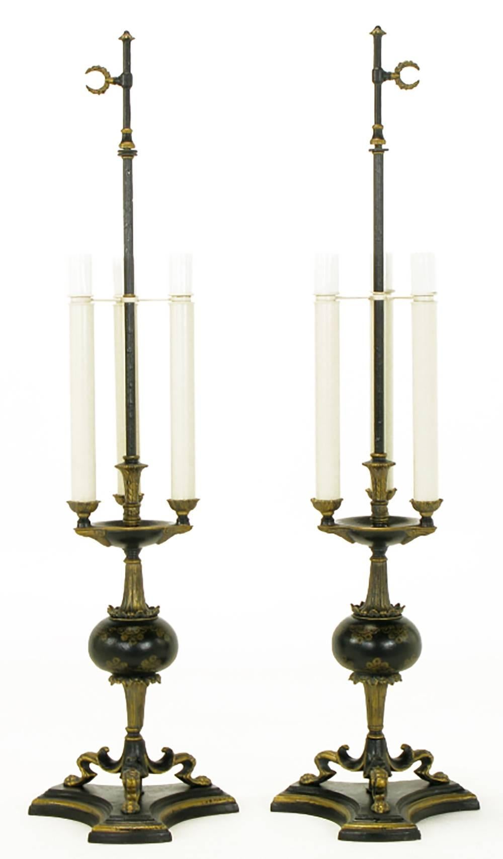 Extremely well-constructed Empire style table lamps of black lacquered metal with parcel-gilt detailing. Triple lions paw over reverse trefoil base. Centre oval with fluted trumpeted spacers. Tall triple candles with standard sockets. Centre post