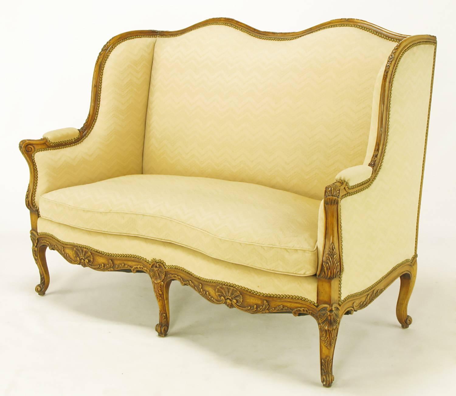 A finely crafted reproduction of an 18th century French settee, this lovely piece features an intricately carved walnut wood frame. Acanthus leaf decoration adorns the wooden arms and cabriole legs, with carved sea shells and scroll feet adding