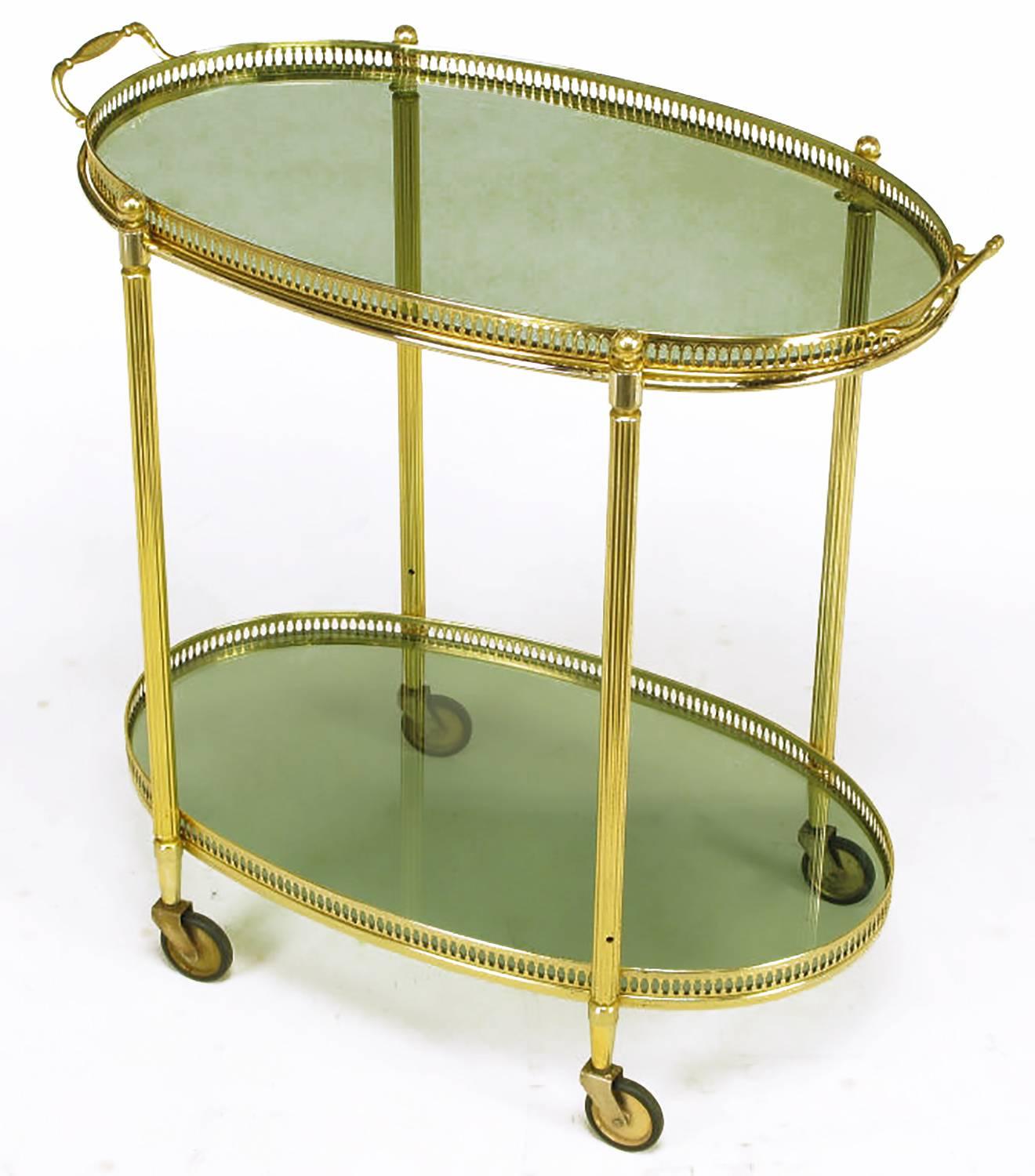 Two-tiered bar cart with pierced brass gallery and reeded brass legs. Oval smoked glass surfaces with removable two-handle top tray. Rubber tires on brass casters.