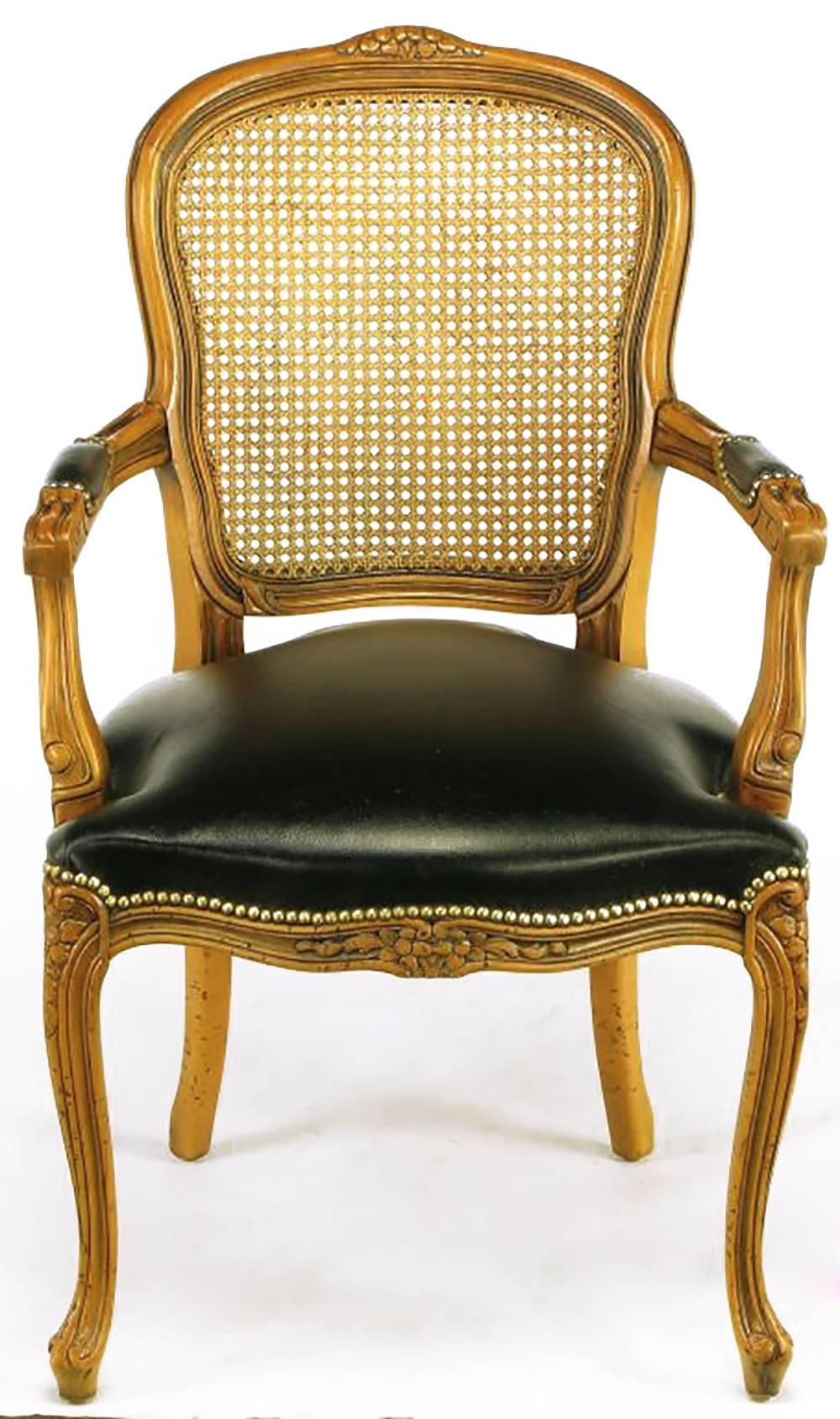 Set of four Louis XV style carved fruitwood armchairs with black leather seats, arm pads and caned backs. Ornately carved apron, turned feet and arms. Finished in a light cherry antiqued glaze with speckled worm hole patina. Brass nail head