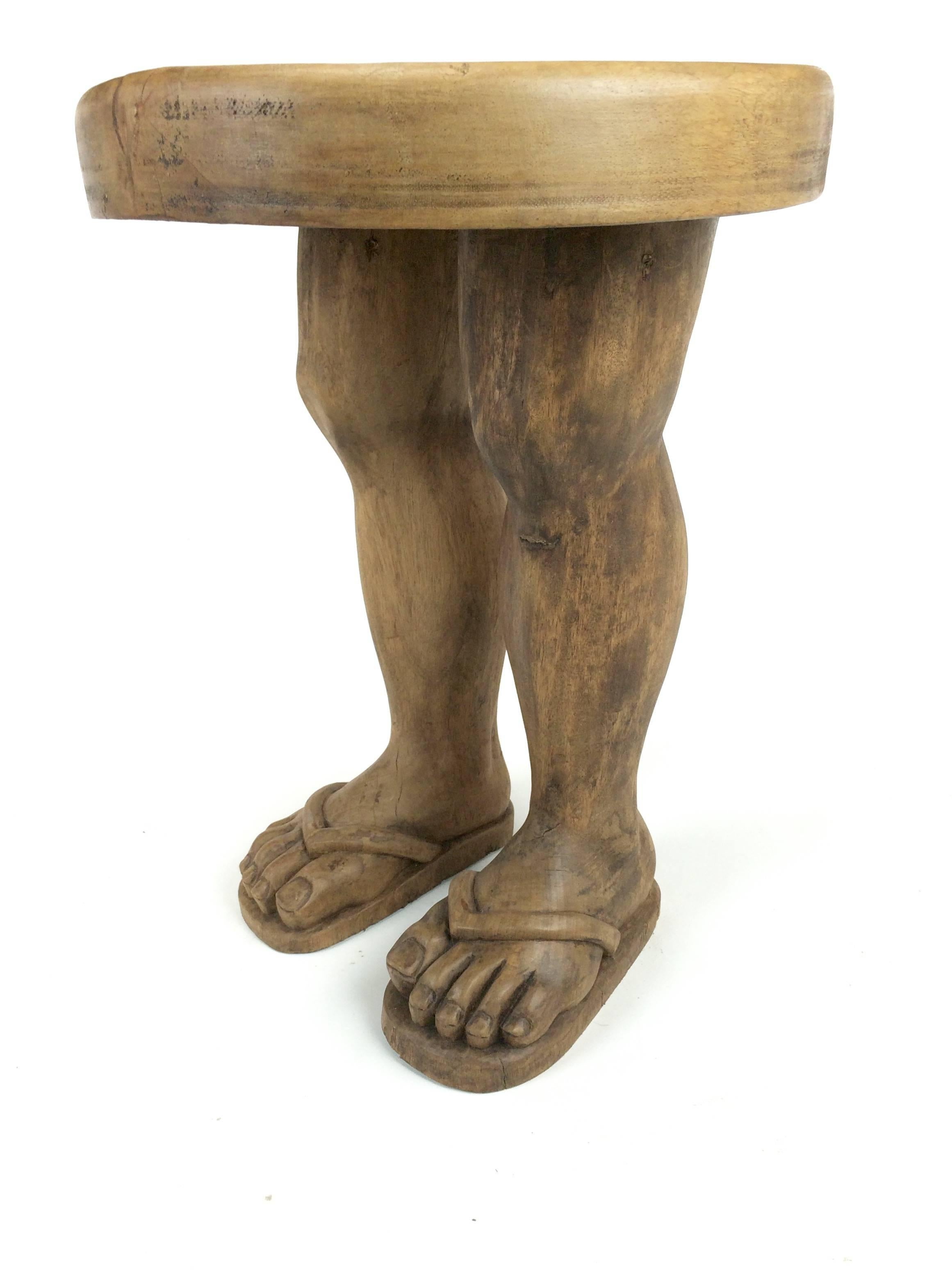 Hand-carved solid wood Folk Art stool in the shape of two legs, sandaled feet and thick wood disc seat.