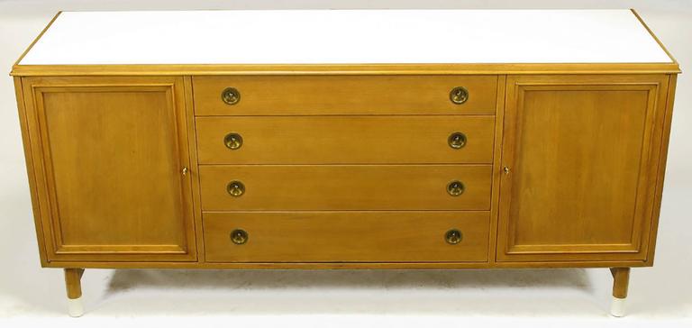 Attributed to Renzo Rutili for Johnson Furniture, long cabinet finished in bleached mahogany with white Micarta top and white resin sabots. Circular recessed brass pulls with center studs open the four drawers. Beveled doors open to reveal single
