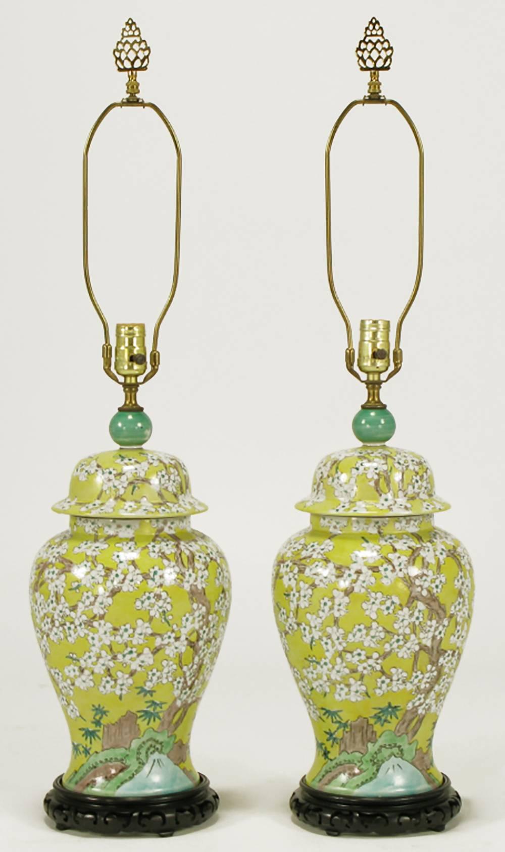 Pair of Chinese import hand-painted yellow glazed ceramic ginger jars with green finial table lamps on carved and ebonized wood bases. Hand-painted white cherry blossoms on branches with a stream, castle and foliate detail. Brass stem, harp, socket