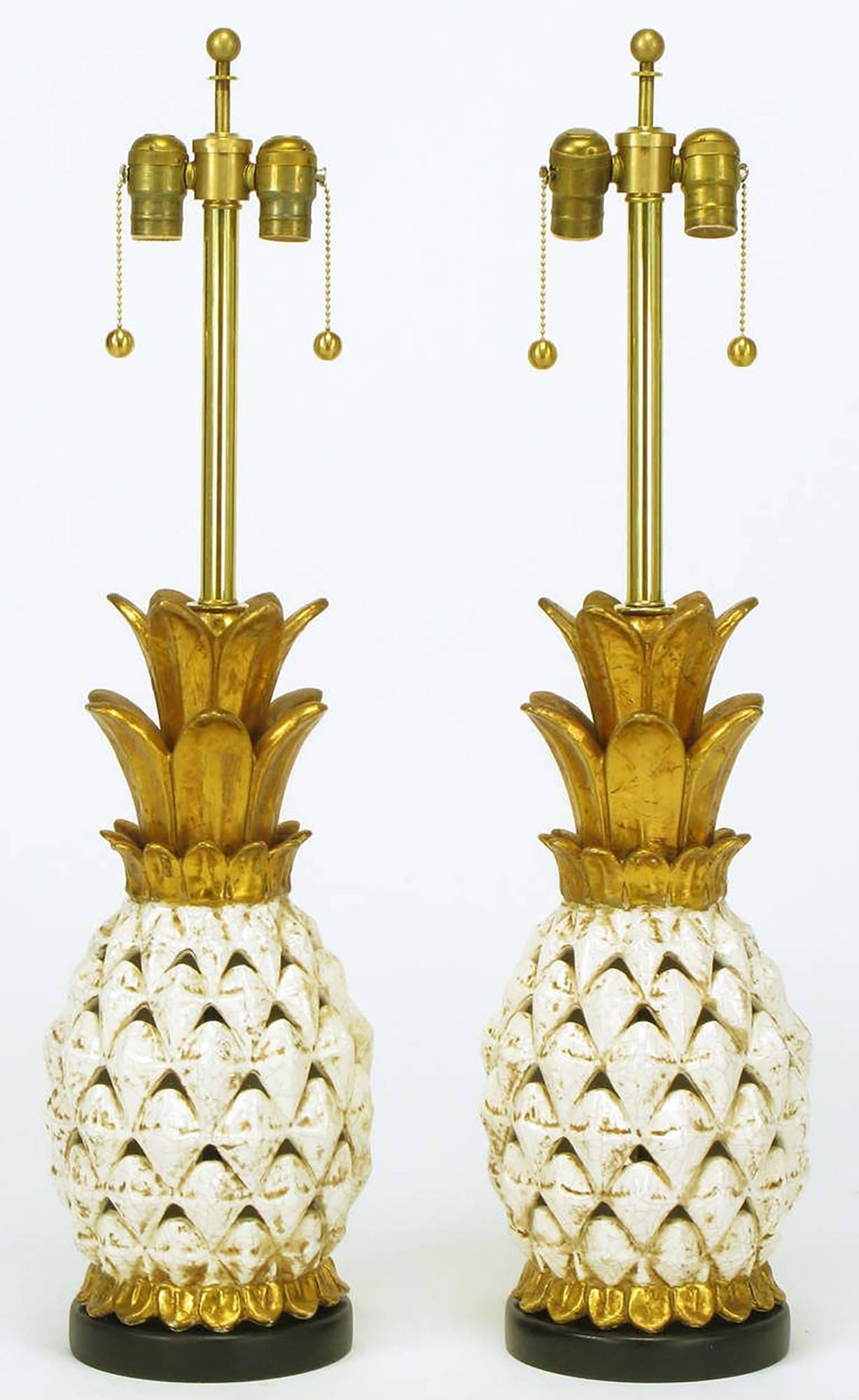 Pair of pierced ceramic body table lamps in the shape of white glazed and parcel-gilt pineapples with gilt crown. Black lacquered wood base, brass stems and double sockets with ball end pull chain.
