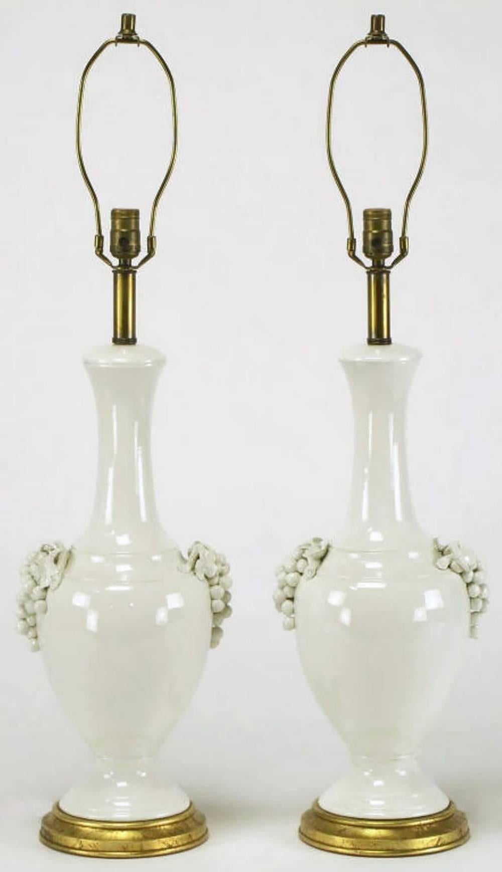 Elegant and early pair of urn form white ceramic table lamps with grape cluster handles from Frederick Cooper Studios. Gilt base with brass stem, socket and harp. Sold sans shades.