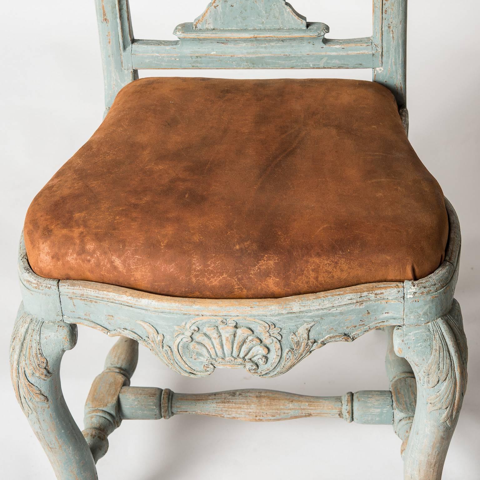 These rare chairs have great form with a fan shaped carving on the crest and old leather seats. The apron echoes the same fan shape as the crest and the legs have intricate carvings at the top and bottom, which is unusual. The old blue paint is a