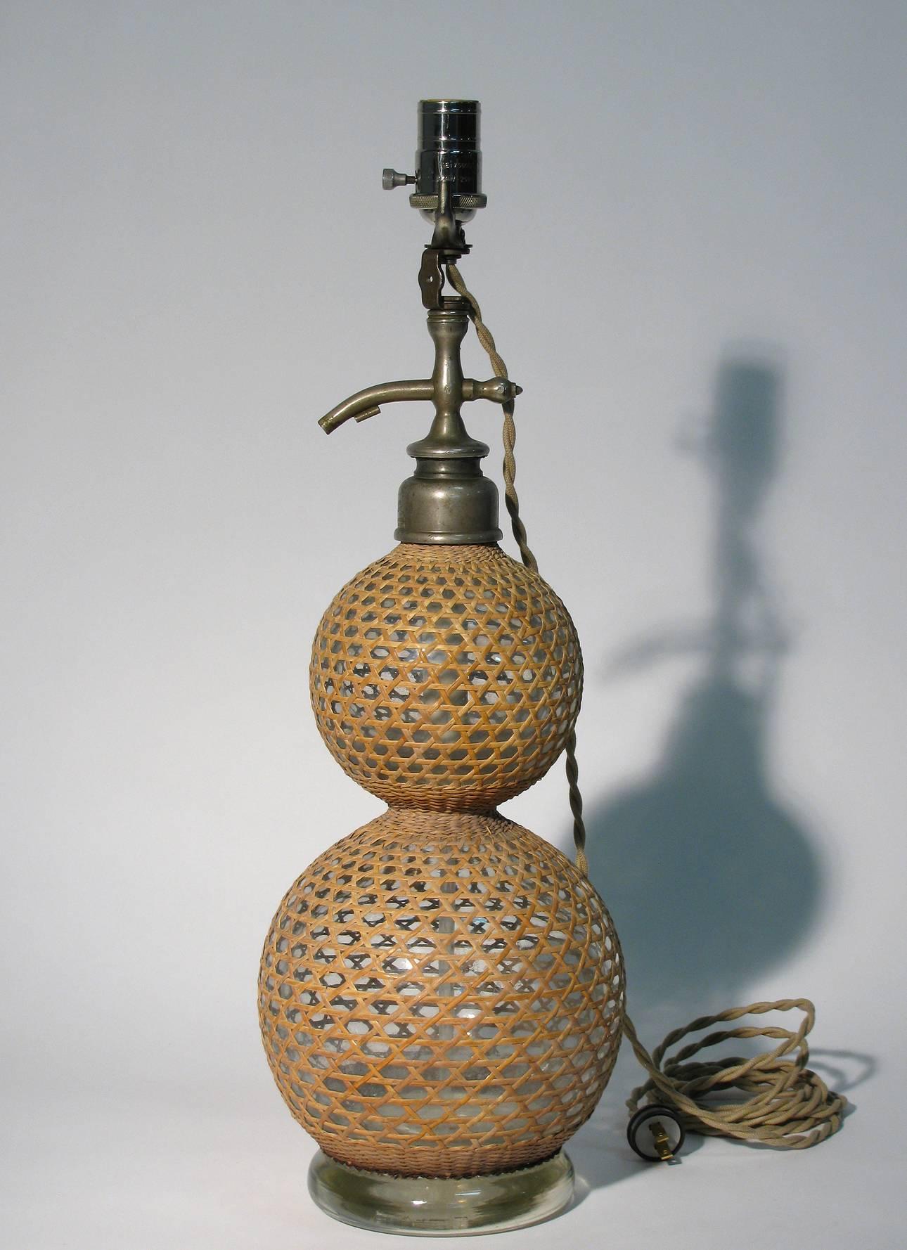 A large cane-wrapped glass seltzer bottle as a table lamp. Signed with hallmark at top, London. Newly wired with tan khaki fabric cord with a single three-way socket. Dimensions: 32 inches height to top of finial, 24 inches to the top of socket, 8