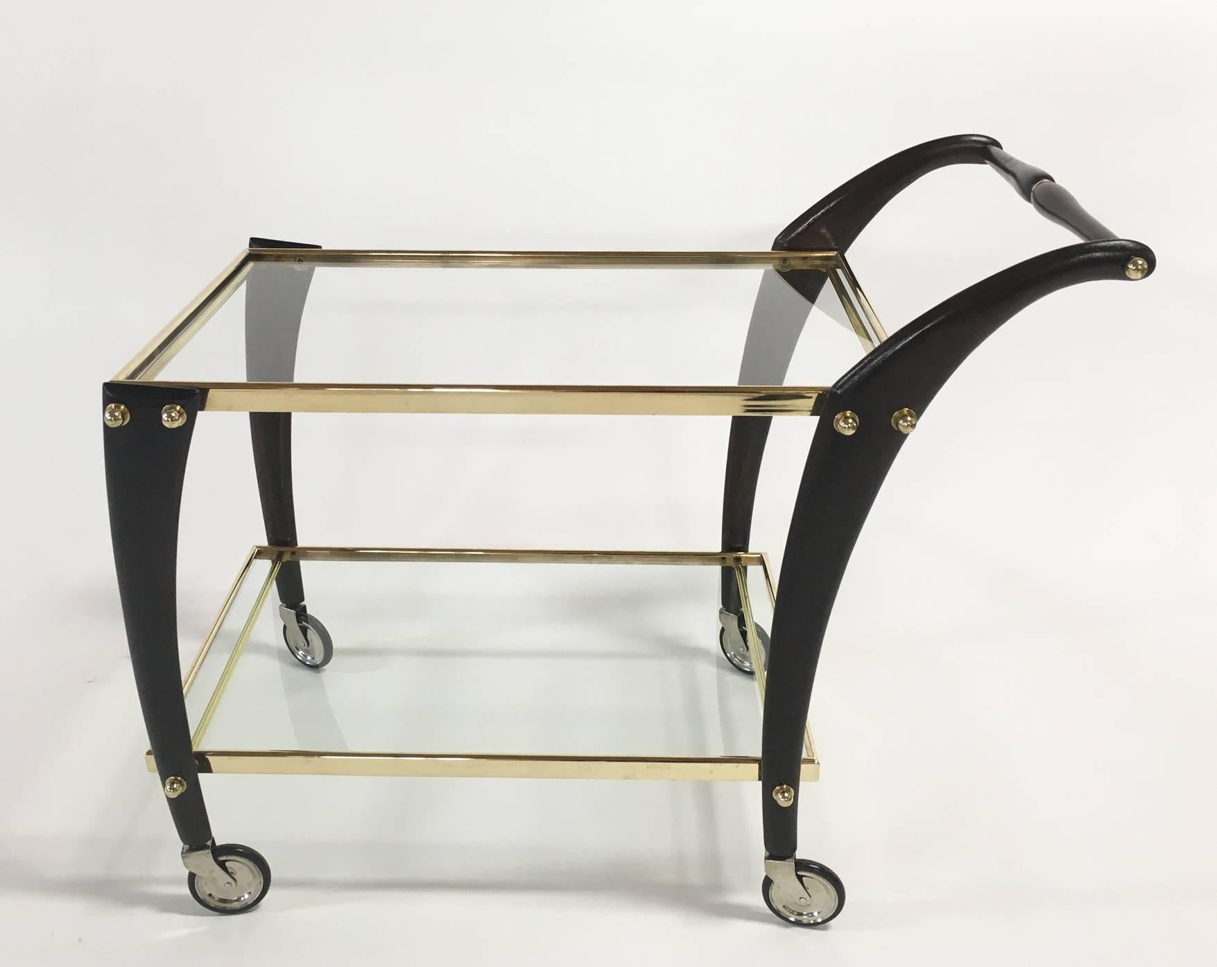 Sculptural bar cart attributed to Cesare Lacca. Two-tiered polished brass frame with clear glass shelves, framed by curvaceous ebonized mahogany posts, on four chrome and rubber swivel wheels. Height measurement with top handle: 27.5 inches.