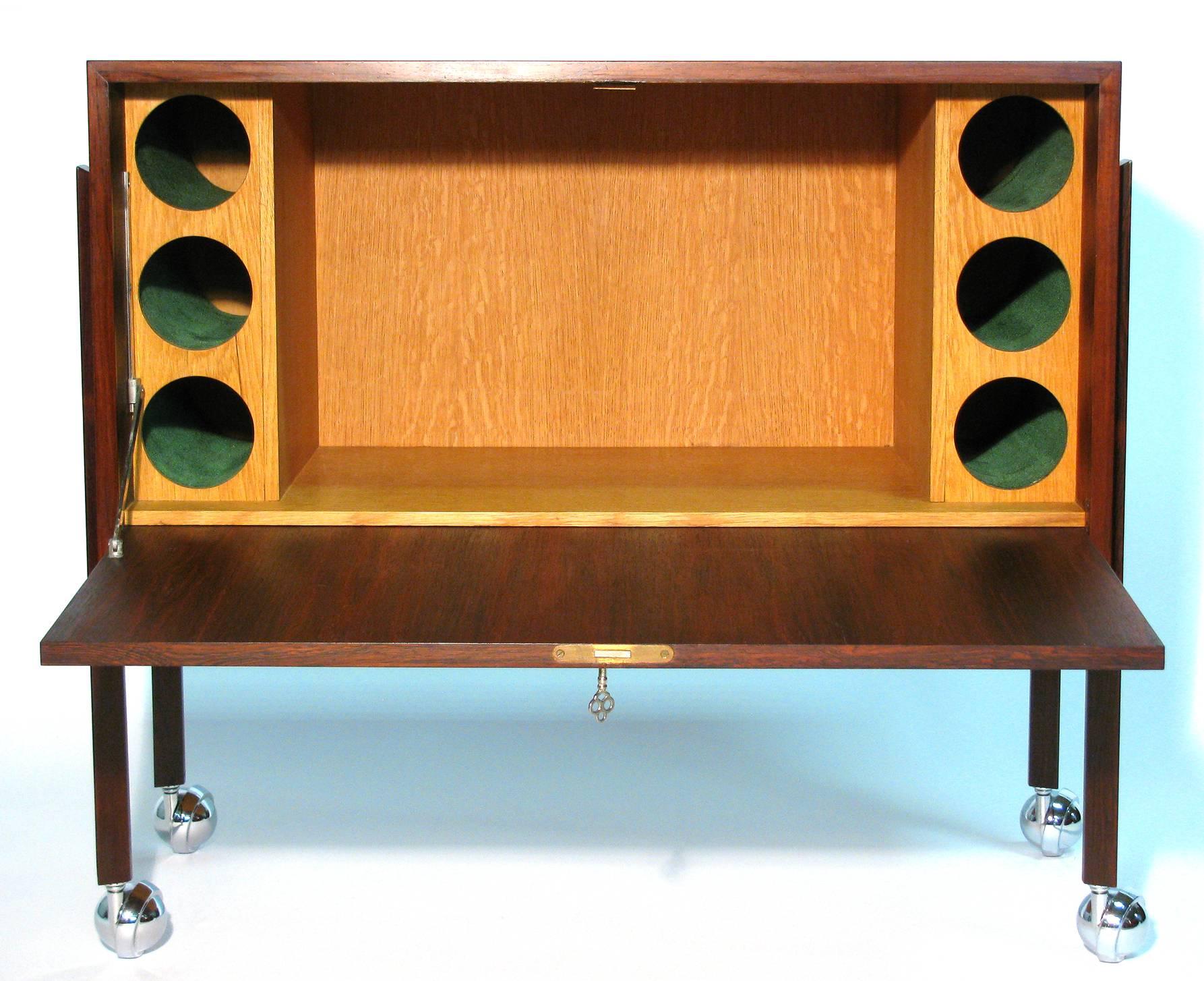 A modernist design liquor cabinet with exceptional book-matched rosewood. Rectangular in form case, suspended by four mounted triangular post legs on chrome ball wheels. The locking drop front opens to reveal a fitted oak interior for storing bar