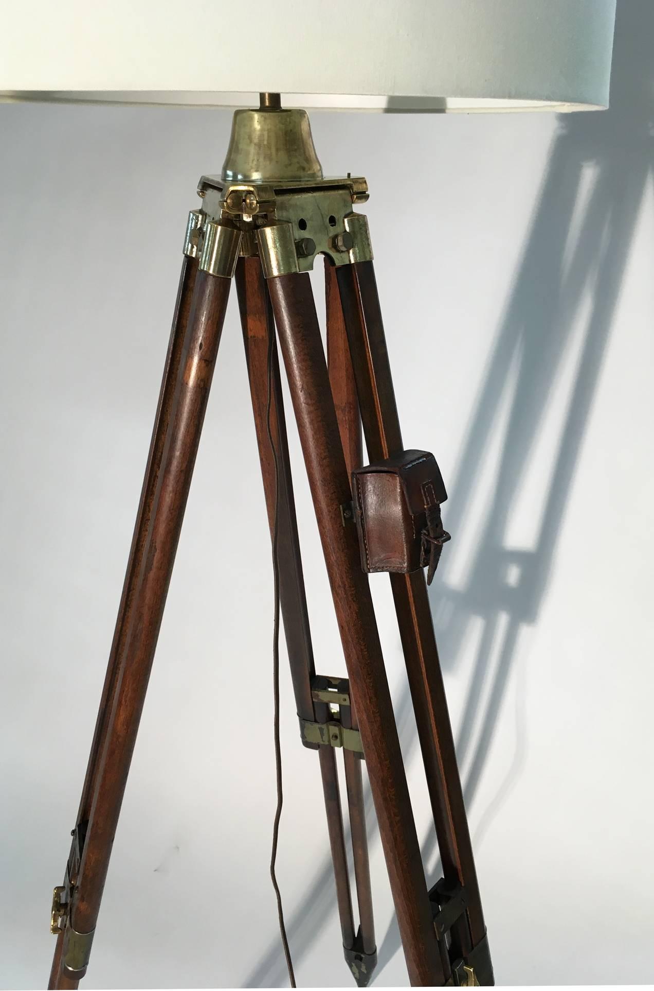 An adjustable floor lamp made from an antique Swiss surveyor tripod of wood and solid brass hardware. Features include brass adjustment wing nuts on patina metal clasps for height adjustment, a small leather box pouch. The tripod base is capped with
