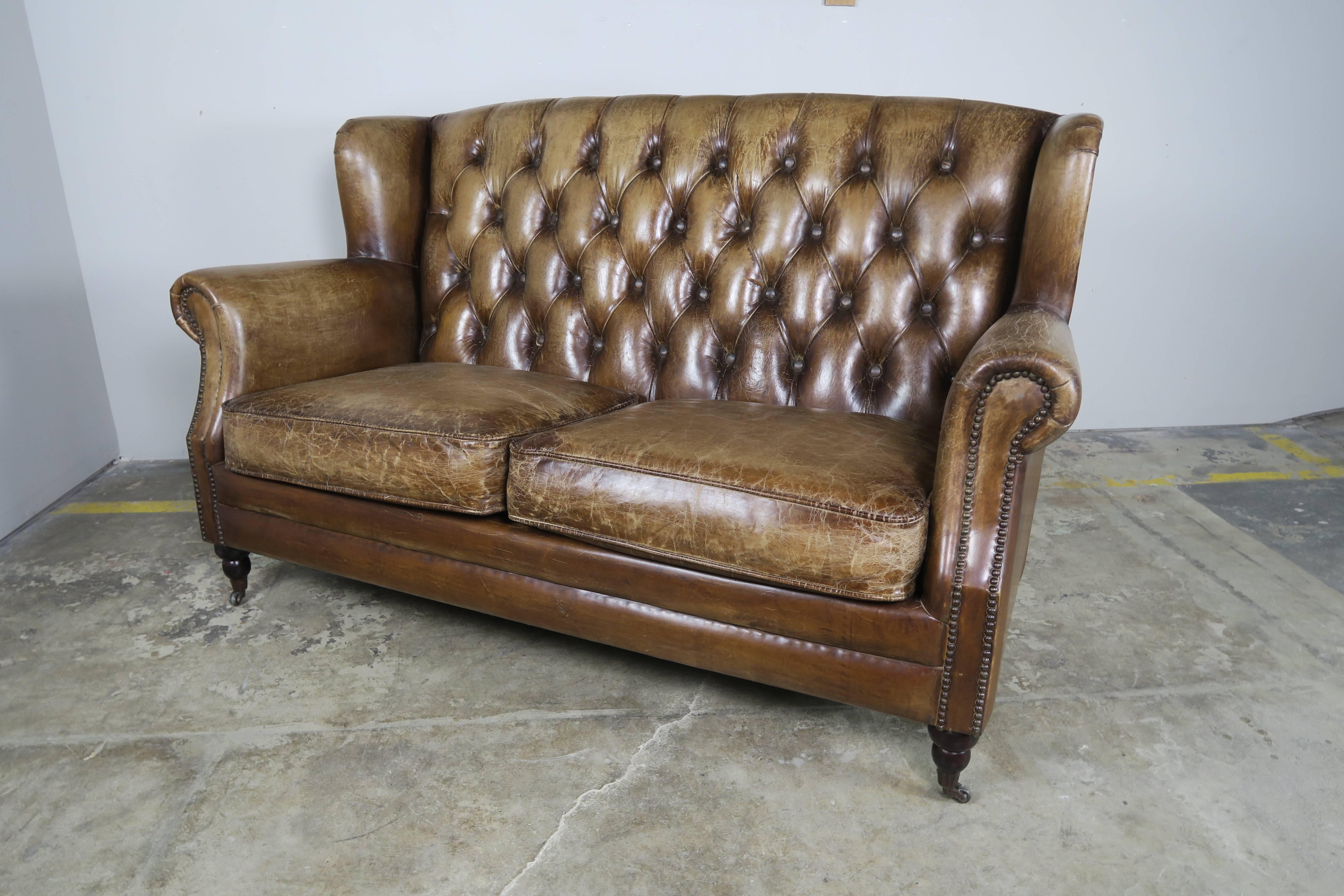 English leather upholstered Chesterfield style sofa standing on four caster legs. Tufted leather back and two loose seat cushions. Nailhead trim detail.