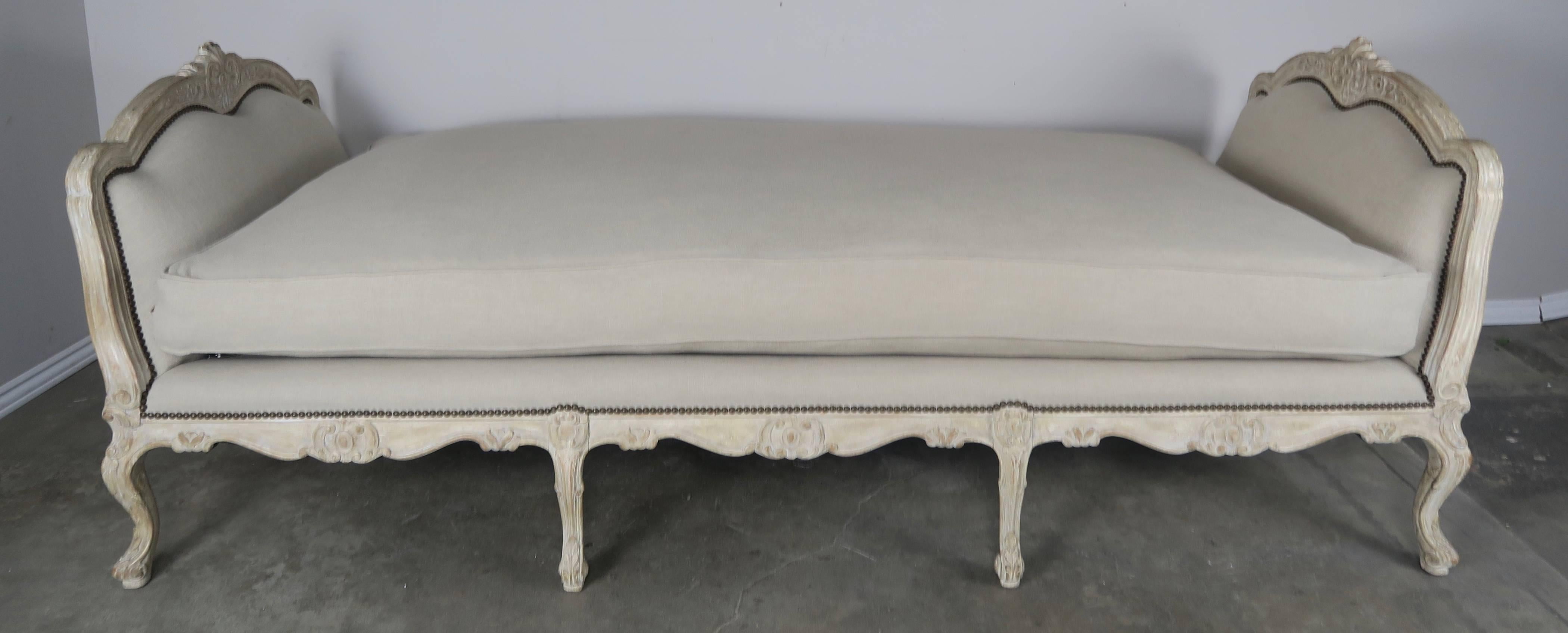 French Louis XV style carved wood daybed in a soft white wash painted finish. The daybed has been newly upholstered in a natural colored linen with antique brass colored nail heads. The daybed stands on eight caved cabriole legs. Down filled