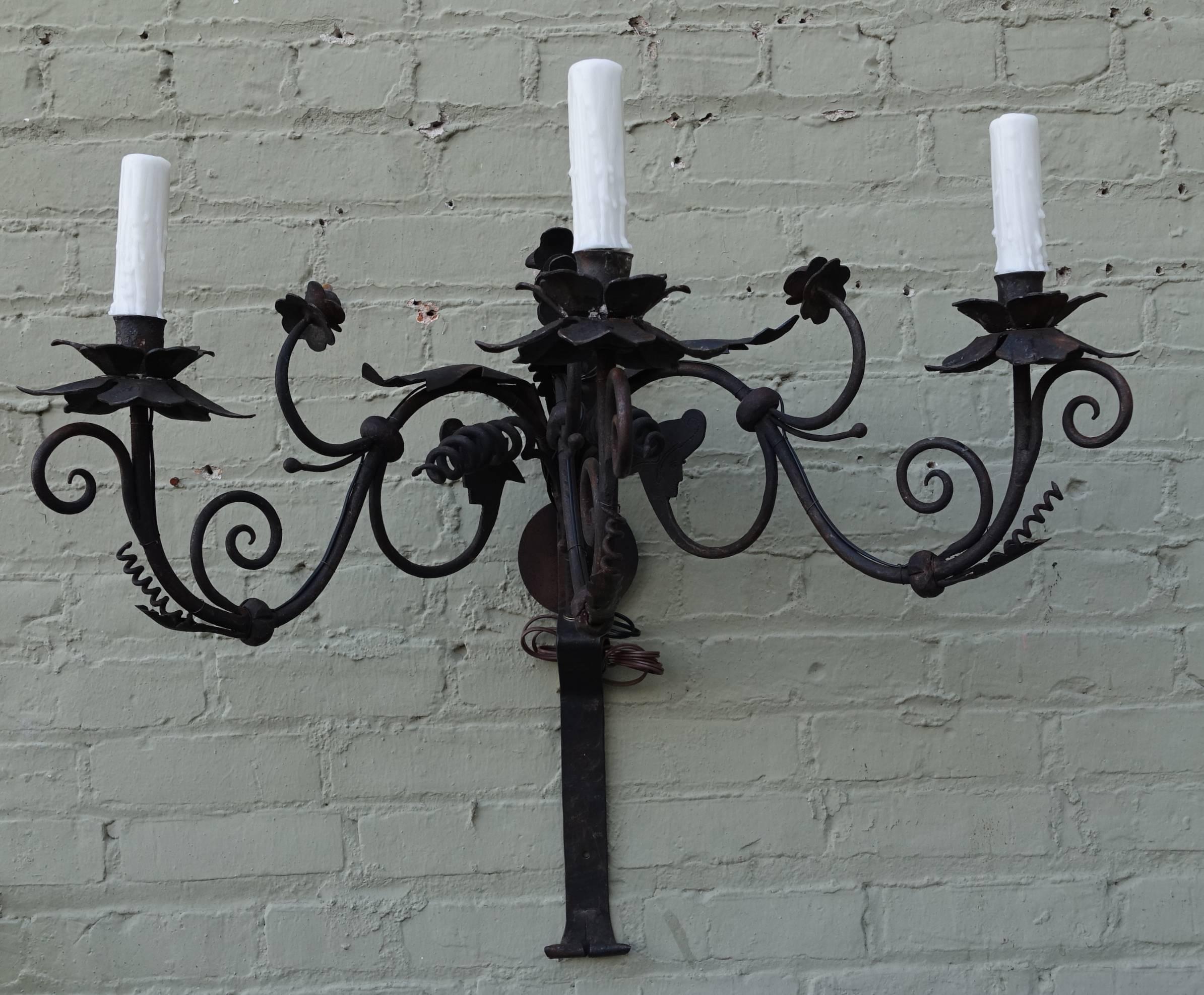 Monumental pair of three-light wrought iron sconces depicting flowers, swirls and faces with wrought iron back plates. The sconces are newly wired with drip-wax candle covers and are ready to install.