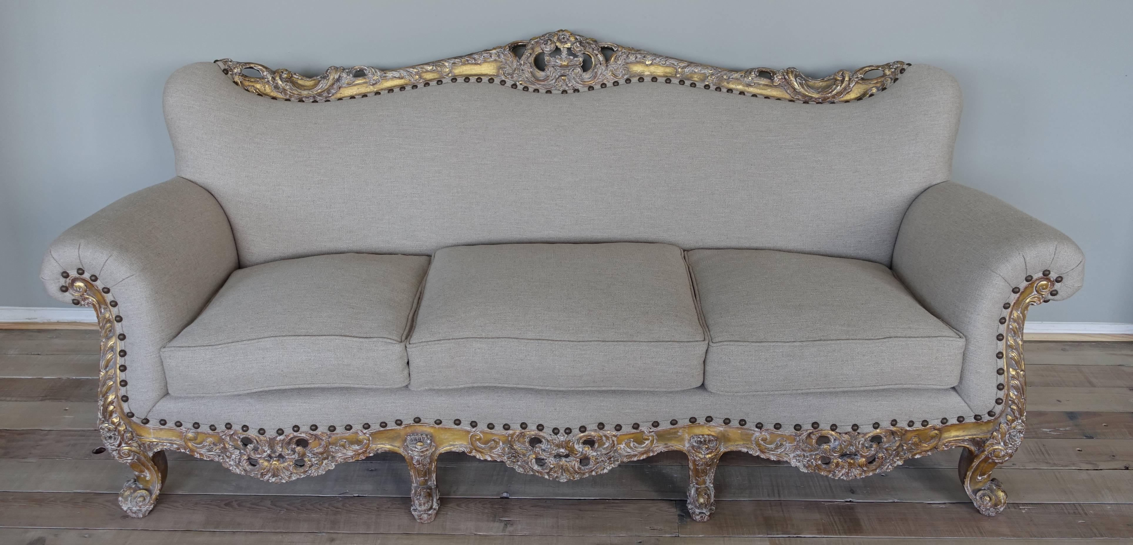 19th century Louis XV style French carved giltwood sofa standing on eight cabriole legs with ram's head feet. Newly upholstered in grain colored natural linen with nailhead trim detail. Carved acanthus leaf detail throughout. Loose down wrapped