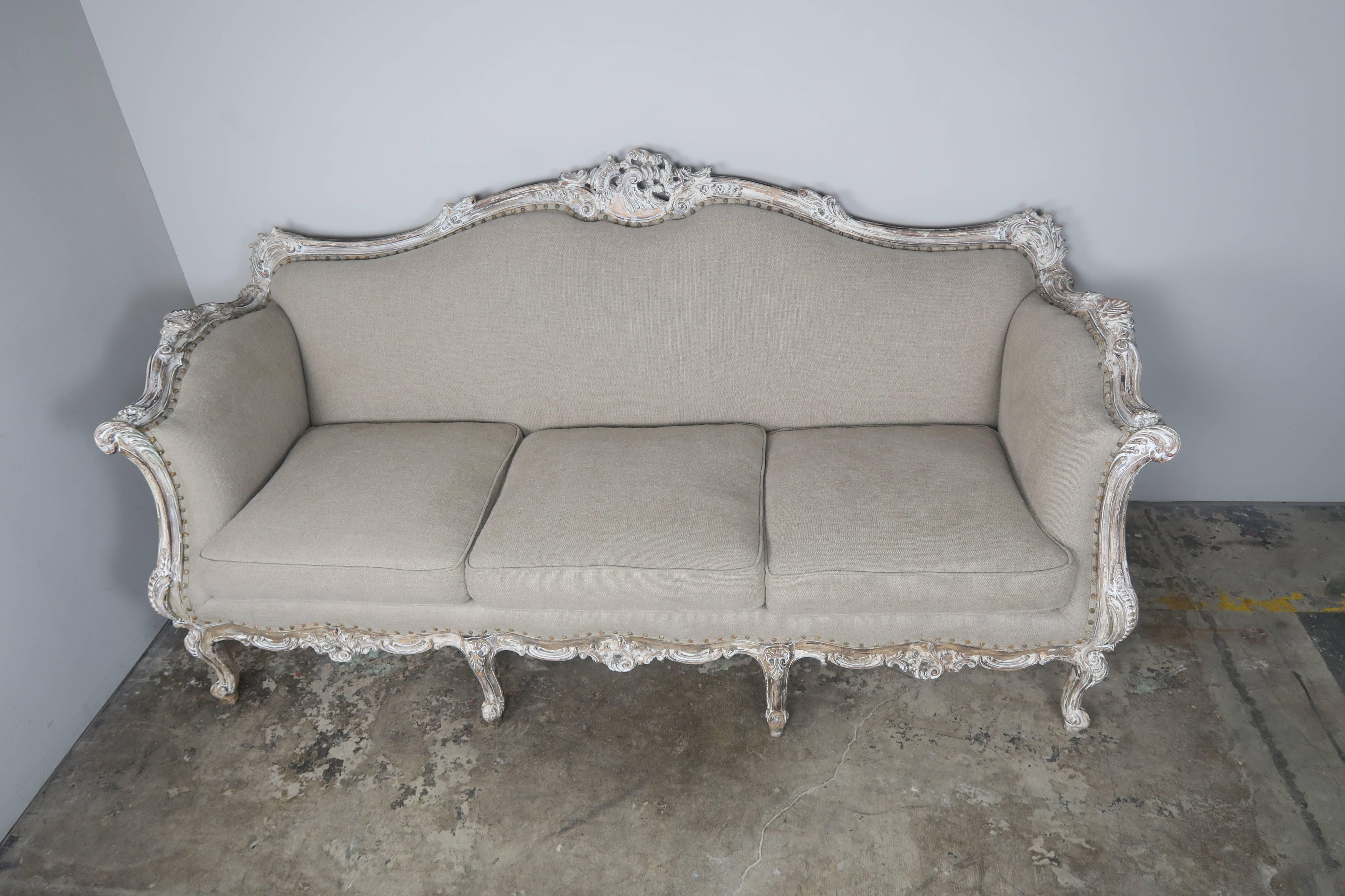 French Rococo style painted sofa standing on eight cabriole legs with ram's head feet. Worn antique white painted finish. The sofa is newly upholstered in a natural Belgium style linen with original nailhead trim detail. Three envelope cushions with