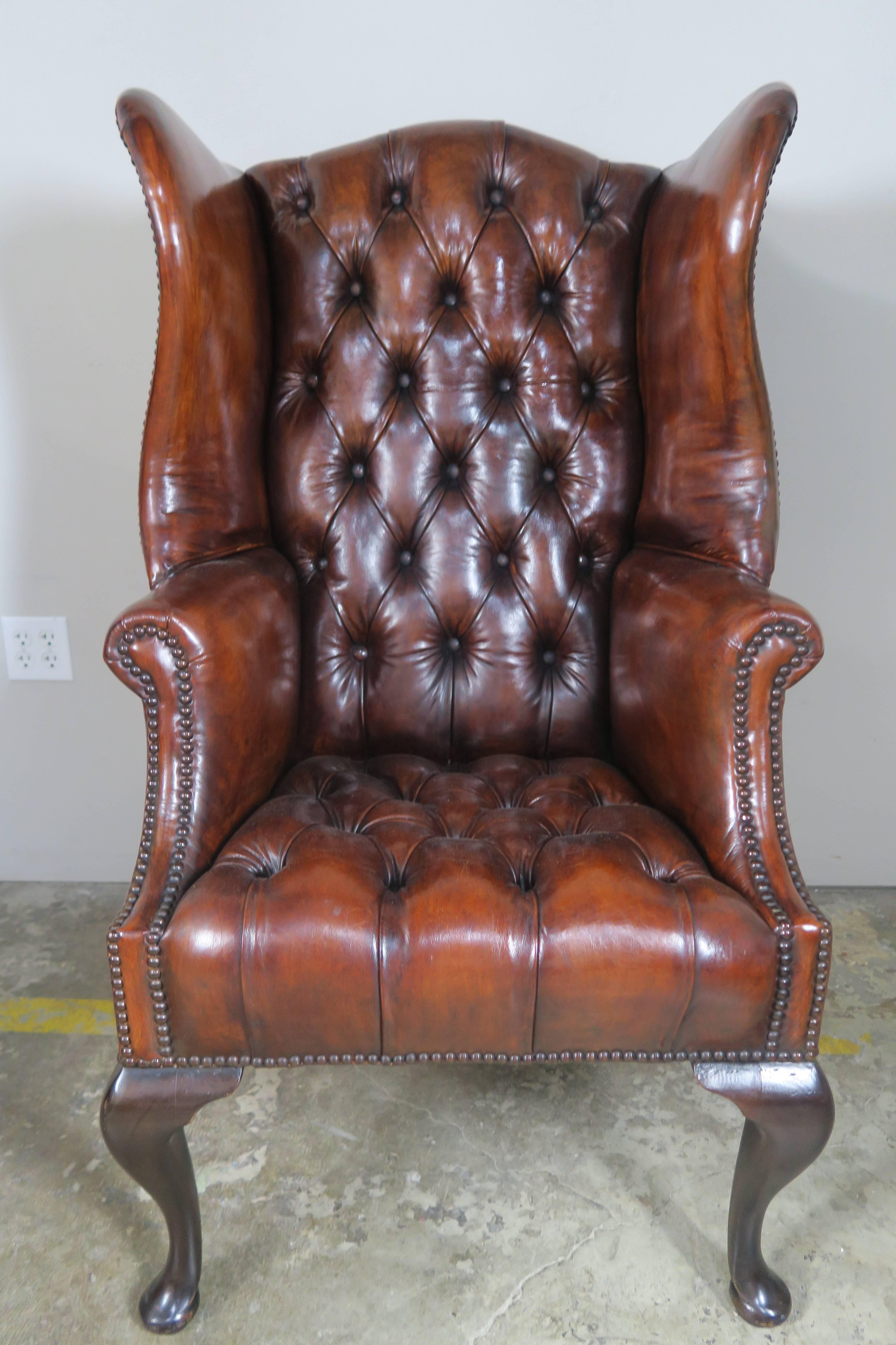 Chestnut colored leather tufted 1900s English wing back armchairs standing on four Queen Anne style legs. Finished with antique brass nailhead trim detail.
 