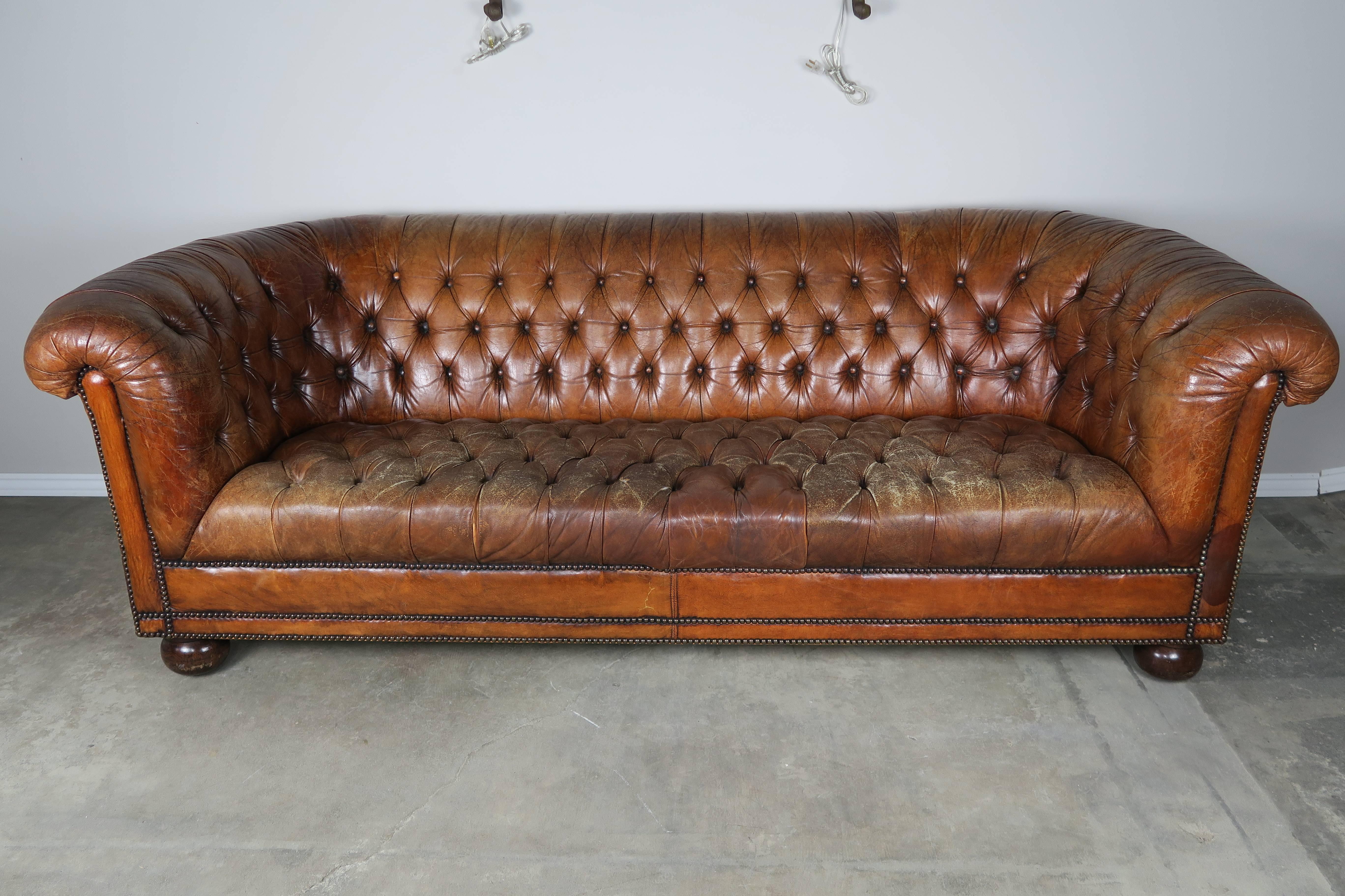English brown leather tufted Chesterfield style sofa standing on four bun feet with nailhead trim detail. Worn, distressed leather consistent with age and use.