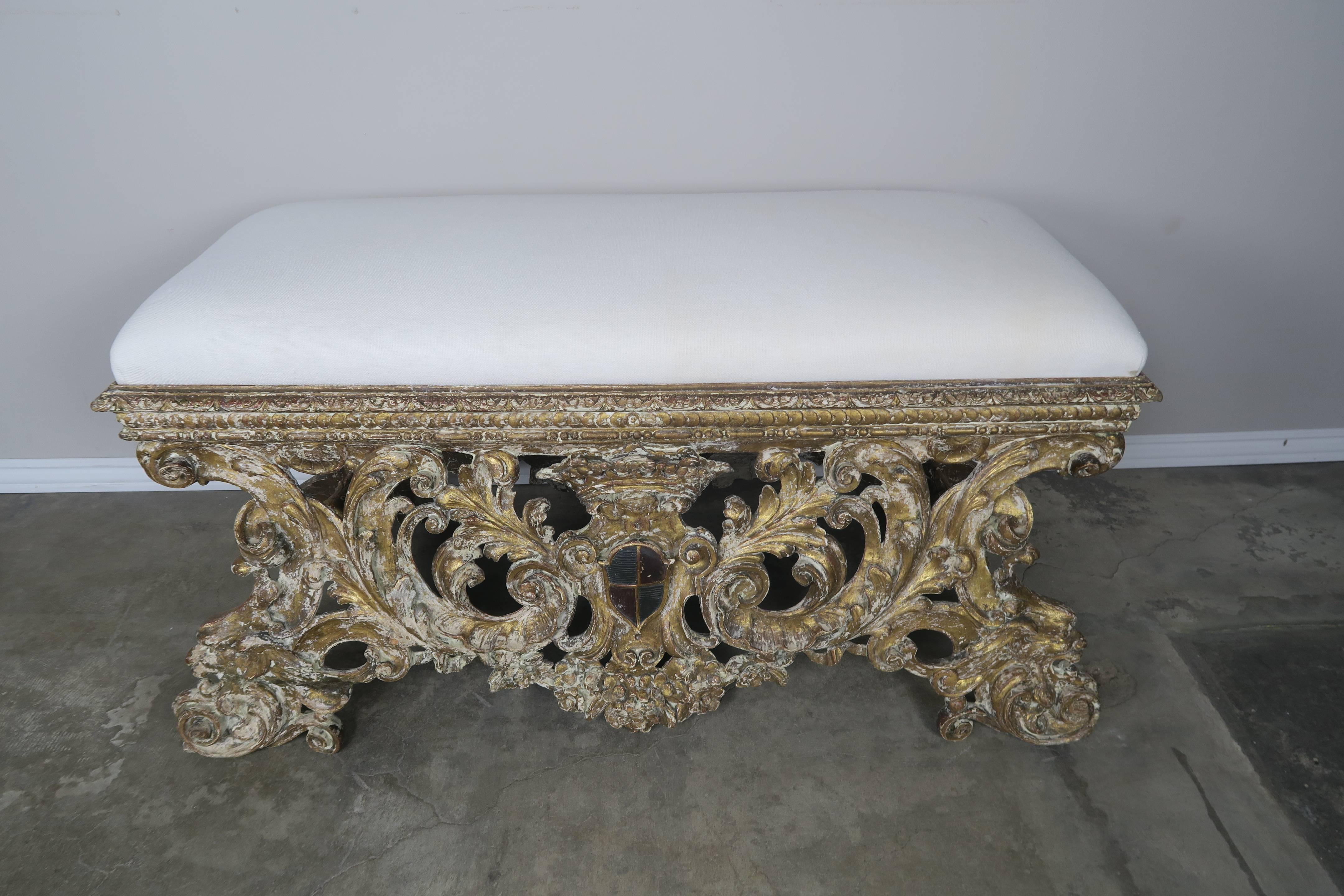 18th century, Italian Baroque style bench carved in wood with swirling acanthus leaves throughout and a center painted coat of arms and crown. Beautiful distressed gold gilt finish. Newly upholstered in a linen textile.
