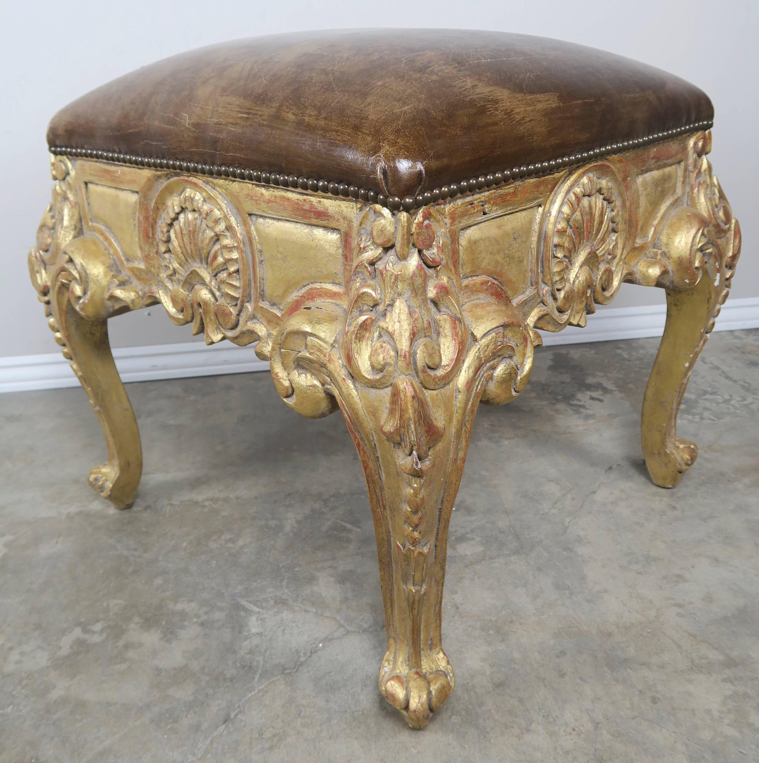 A pair of French 22-karat giltwood carved Louis XV style benches upholstered in beautiful distressed leather with antique brass nailhead trim detail. Carved shell motif on all four sides. The benches stand on cabriole legs that end in ram's head