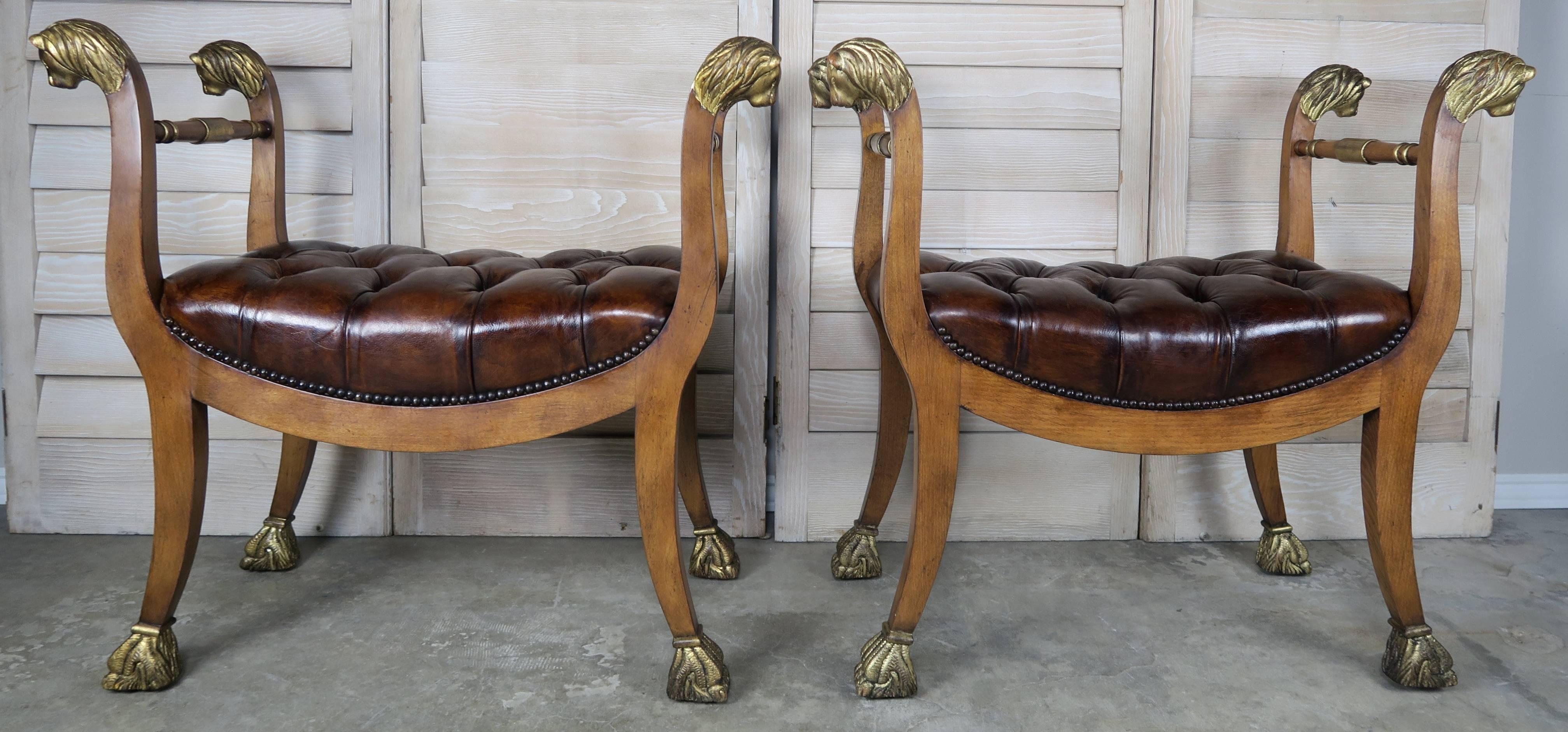Pair of Baker Directoire style gilded 22-karat gold leaf lion head wood stool benches with tufted leather upholstery and nailhead trim detail.