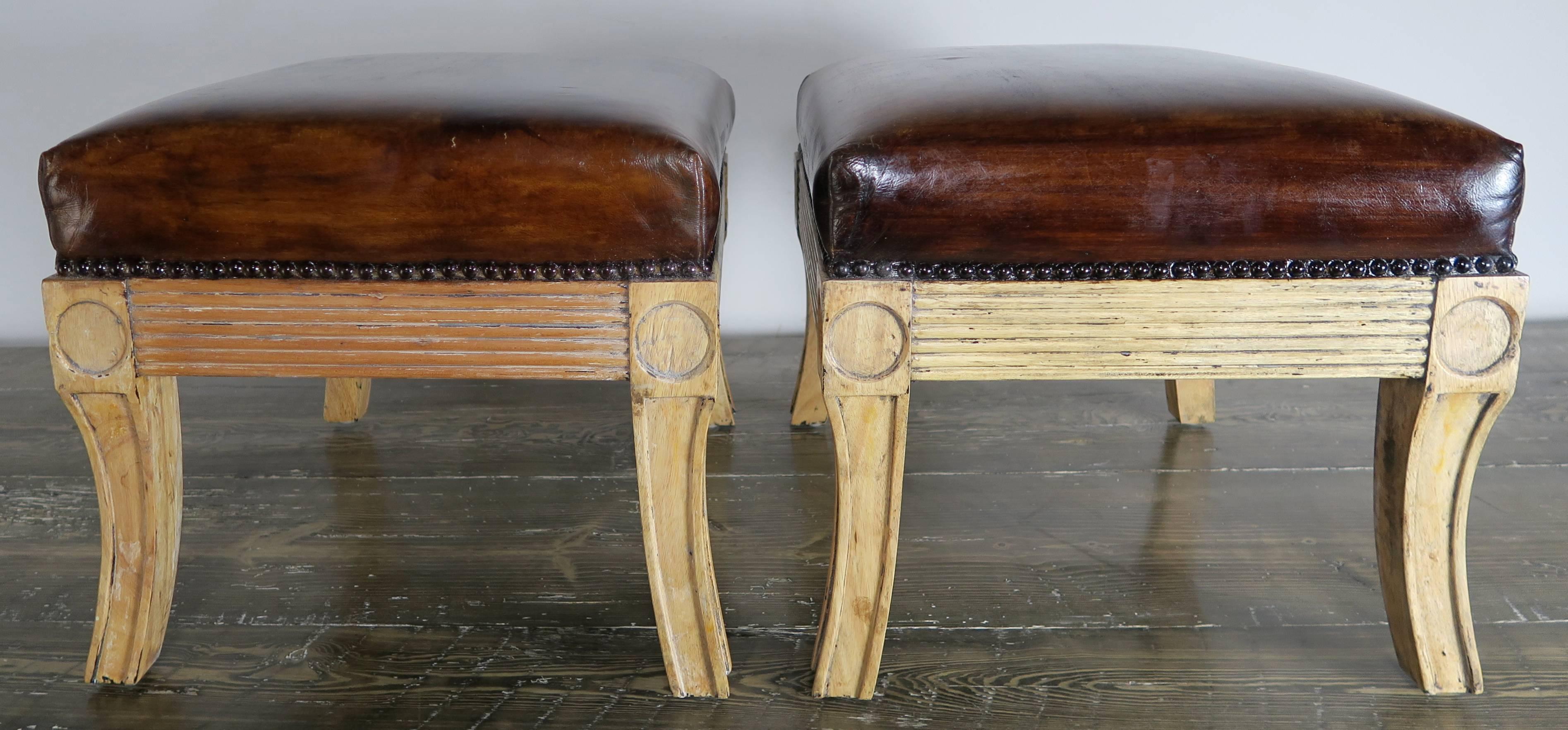 Pair of Italian carved wood benches upholstered in chestnut brown leather with nailhead trim detail. Natural bleached wood finish with remnants of old paint throughout.