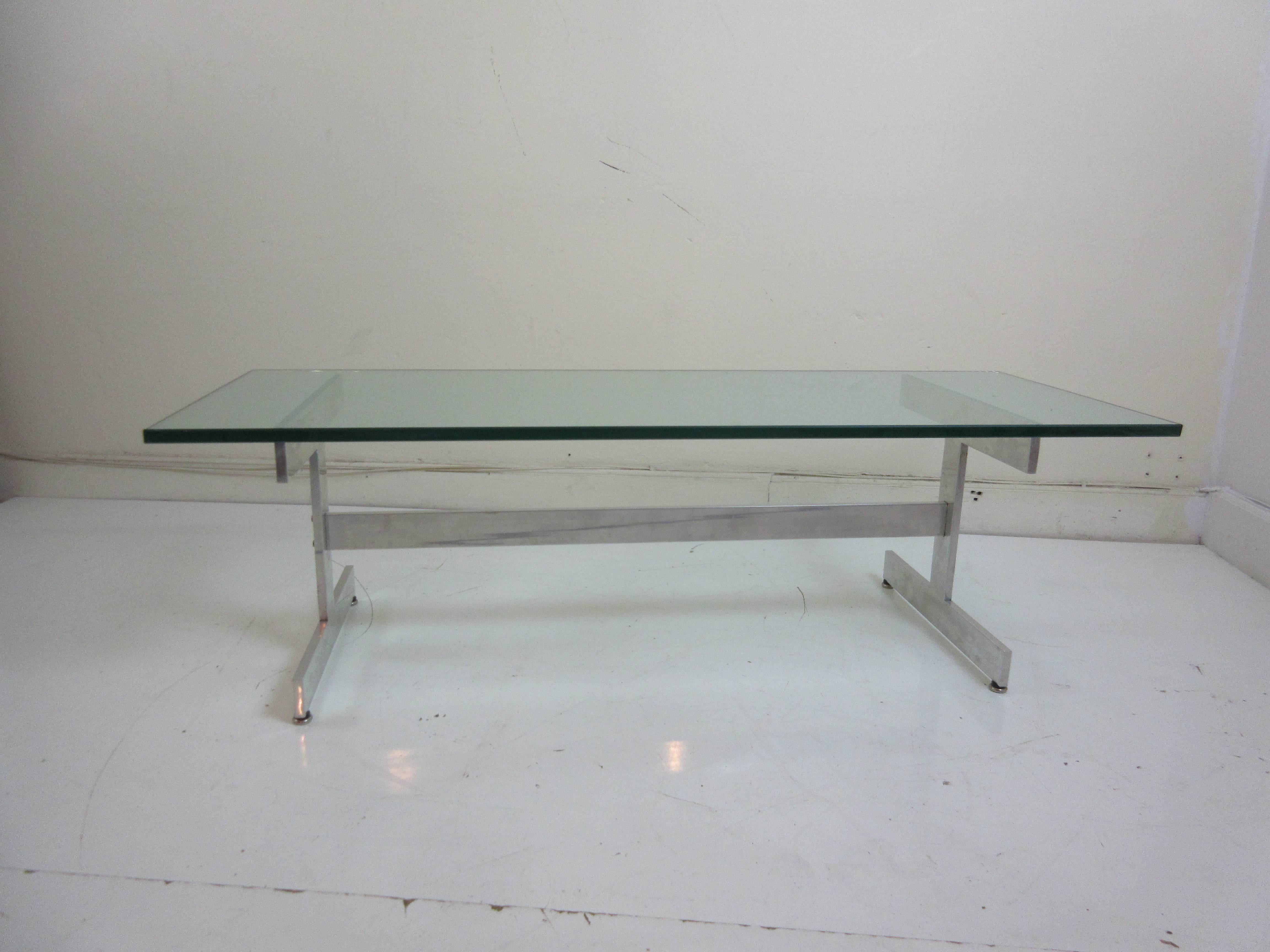 Glass and aluminium coffee table with trestle base connecting H-shaped supports on either side.