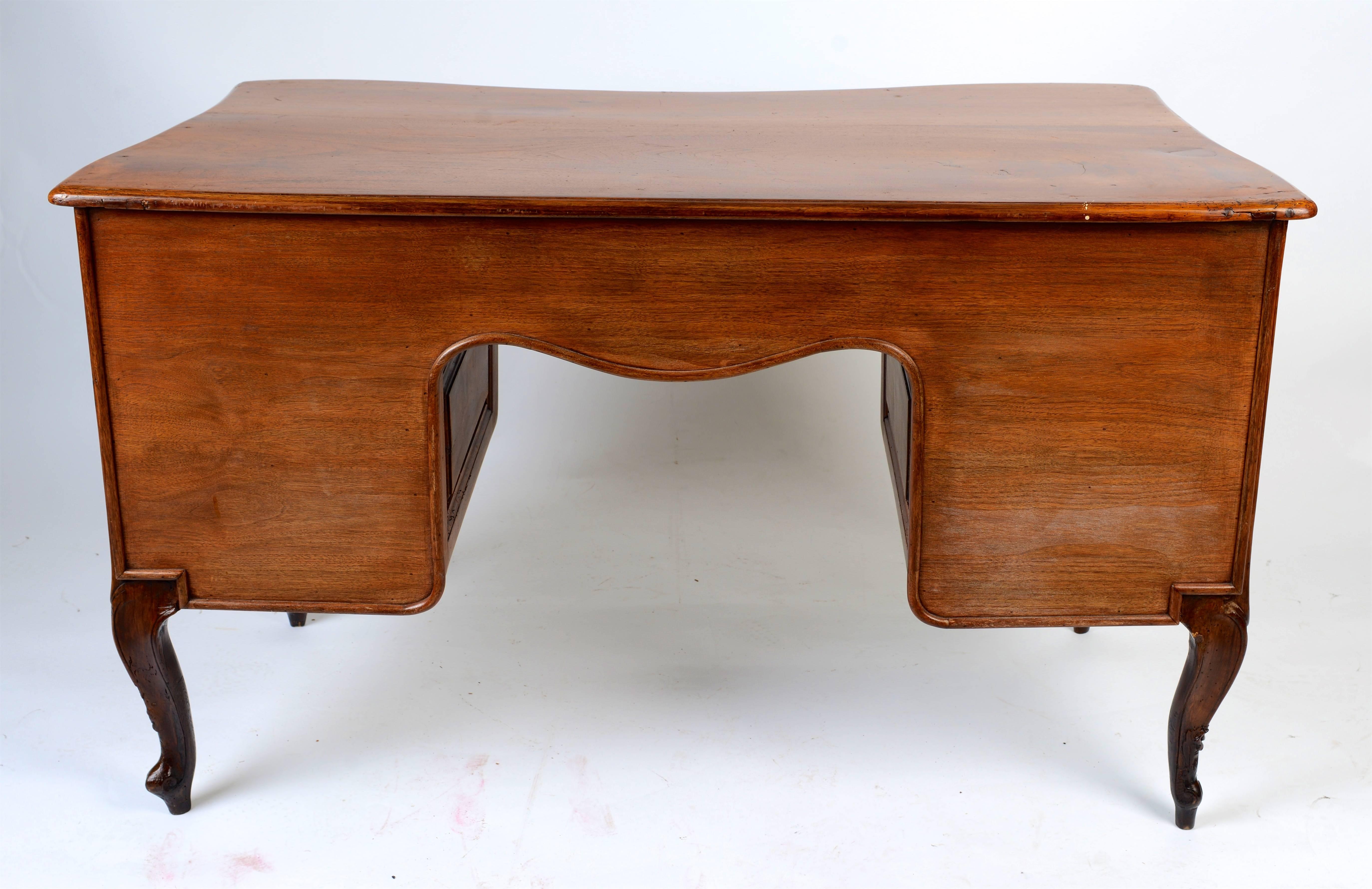 This graceful French desk has a shaped single plank walnut top and a softly curved front with seven curved, raised panel drawers. The knee hole apron is carved with a centre shell surrounded by scrolls that coordinate with the drawer mounts having
