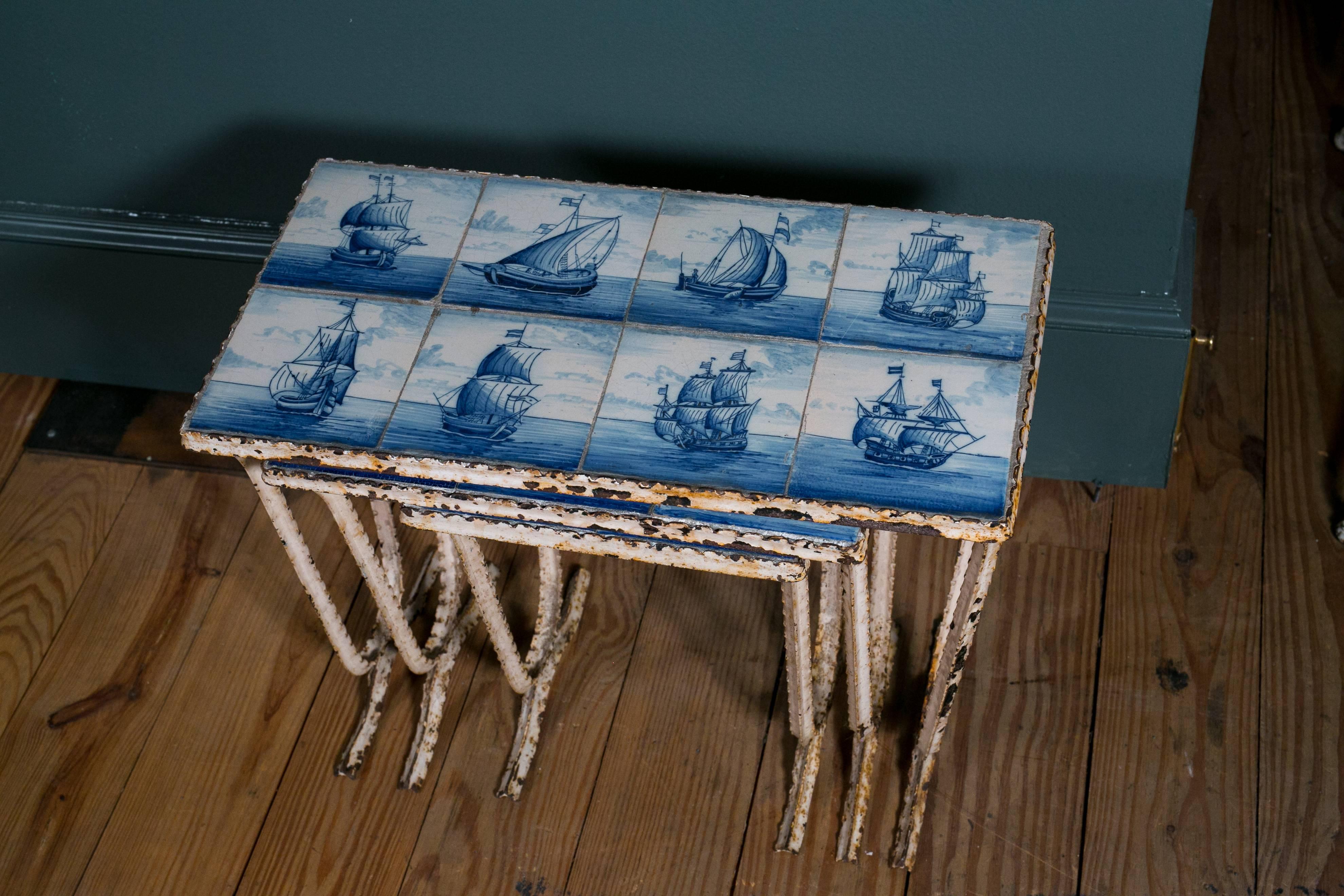 Set of three nesting tables with painted wrought iron bases and Delft blue and white tile tops. Sail boat scenes on each of the tiles. Measurement listed is for largest. Small measures 10.75 inches square x 15.5 inches H. Medium measures 16 inches L