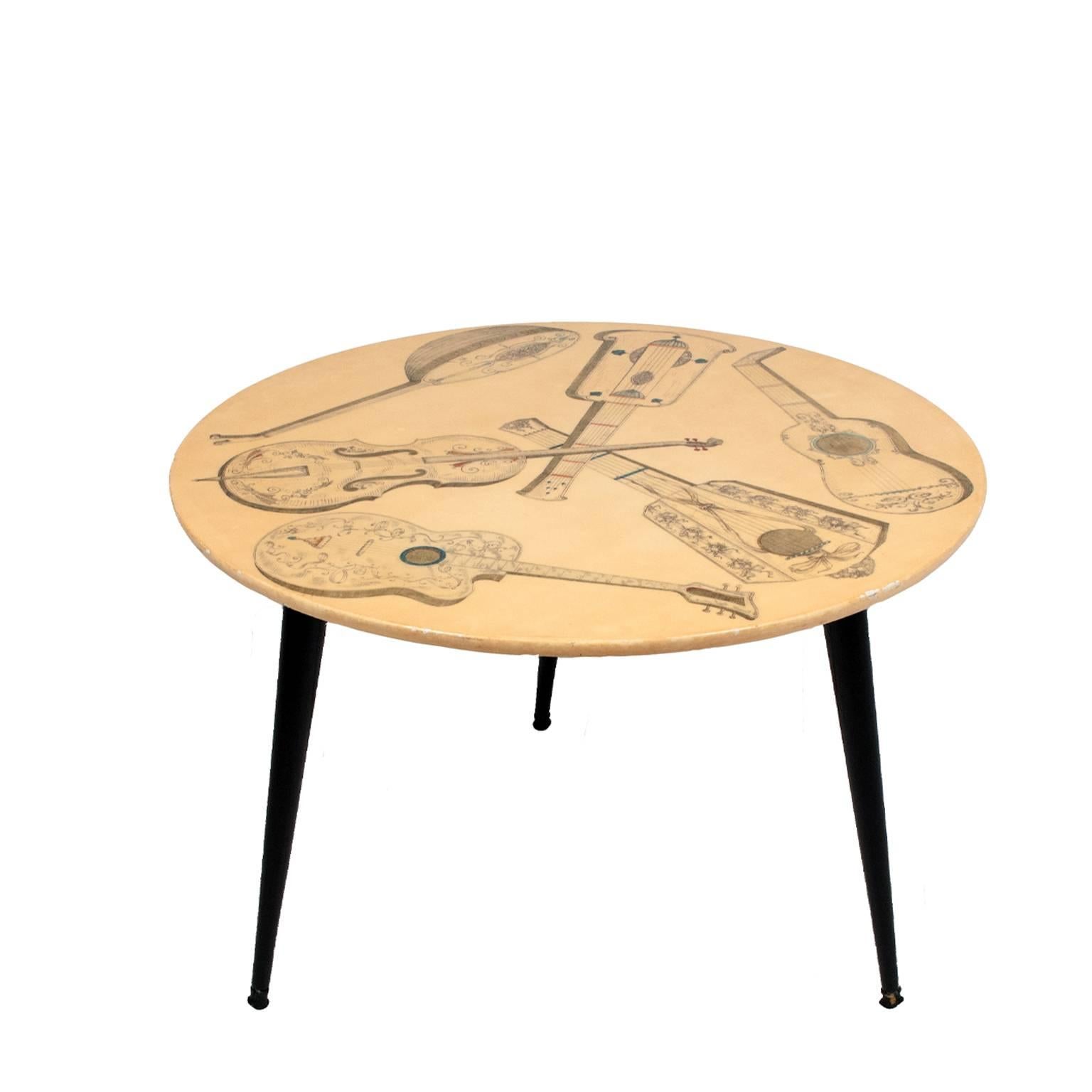 Round top table printed in Fornasetti's signature trompe l'oeil motif; lithographically transfer printed, this table features Strumenti Musicale to the top. Three black lacquered solid wood legs. Label to bottom stamped with number and Italy.