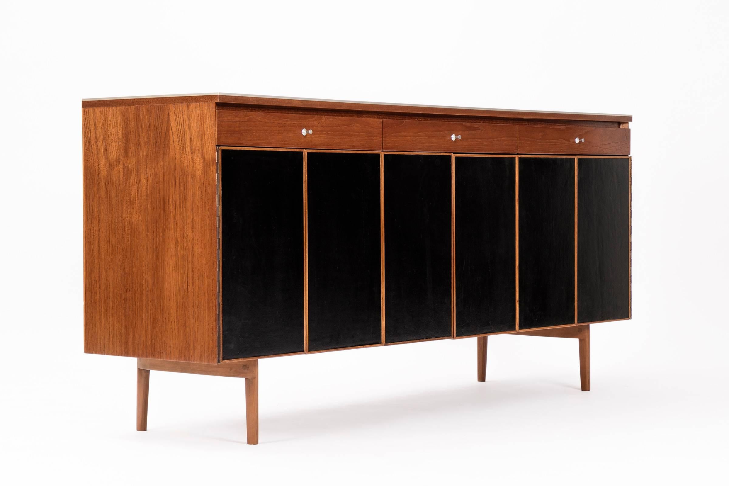 Paul McCobb sideboard features three drawers across the top with aluminium pulls. Piano hinged panels with leather covered fronts conceal six drawers with cut-out tops for pulls.
Walnut interior and exterior have hand rubbed oil finish with solid