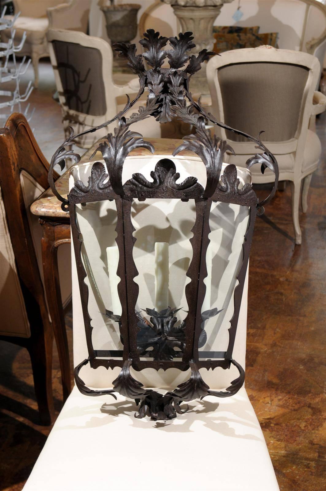 This pair of Italian Rococo style three-light wrought iron wall sconces from the 20th century features exquisite lantern forms. The three authentic wax candle sleeves and their flower shaped iron bobèches are safely protected behind the glass panels