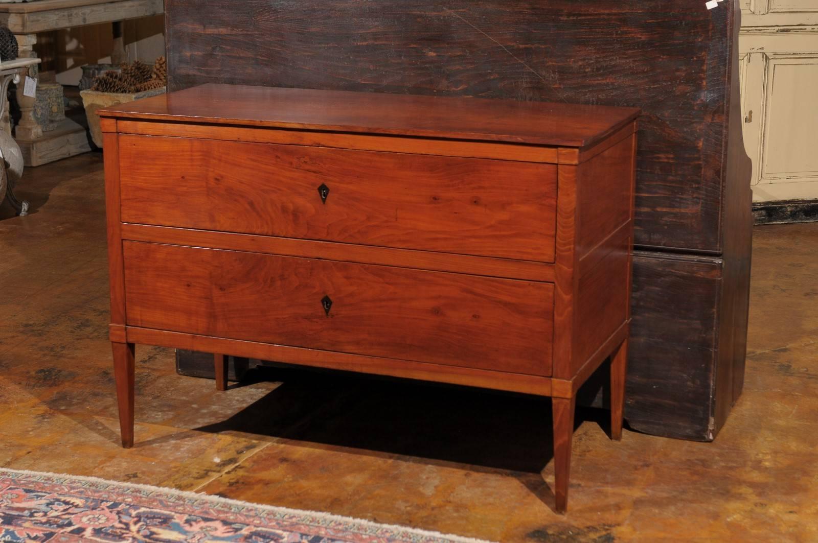 This Italian Neoclassical style fruitwood commode from the late 19th century features a rectangular single plank top over two dovetailed drawers with kite shaped ebonized wood inlaid escutcheons. The sides are divided into two paneled sections. The