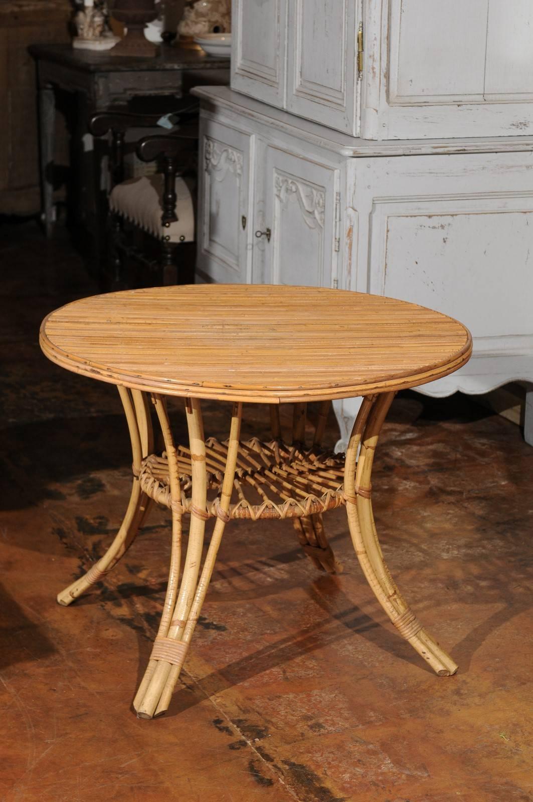 A French mid-century round rattan coffee table with lower shelf and splayed legs. This French rattan side table features a circular top raised over four splayed legs, connected to one another with a smaller pierced shelf. The choice of the simple