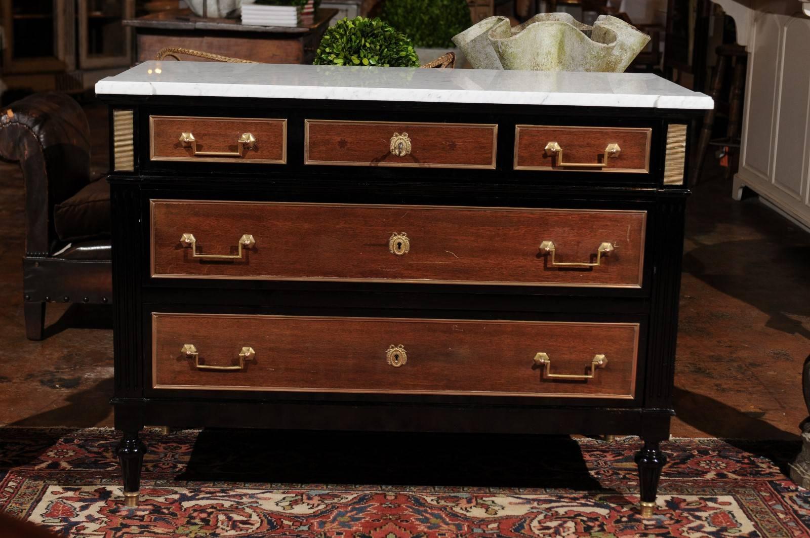 A French Directoire style marble top, ebonized wood with brass accents three-drawer commode from the early 20th century. This exquisite French chest features a white variegated marble top sitting above a frieze drawer. This drawer is divided into