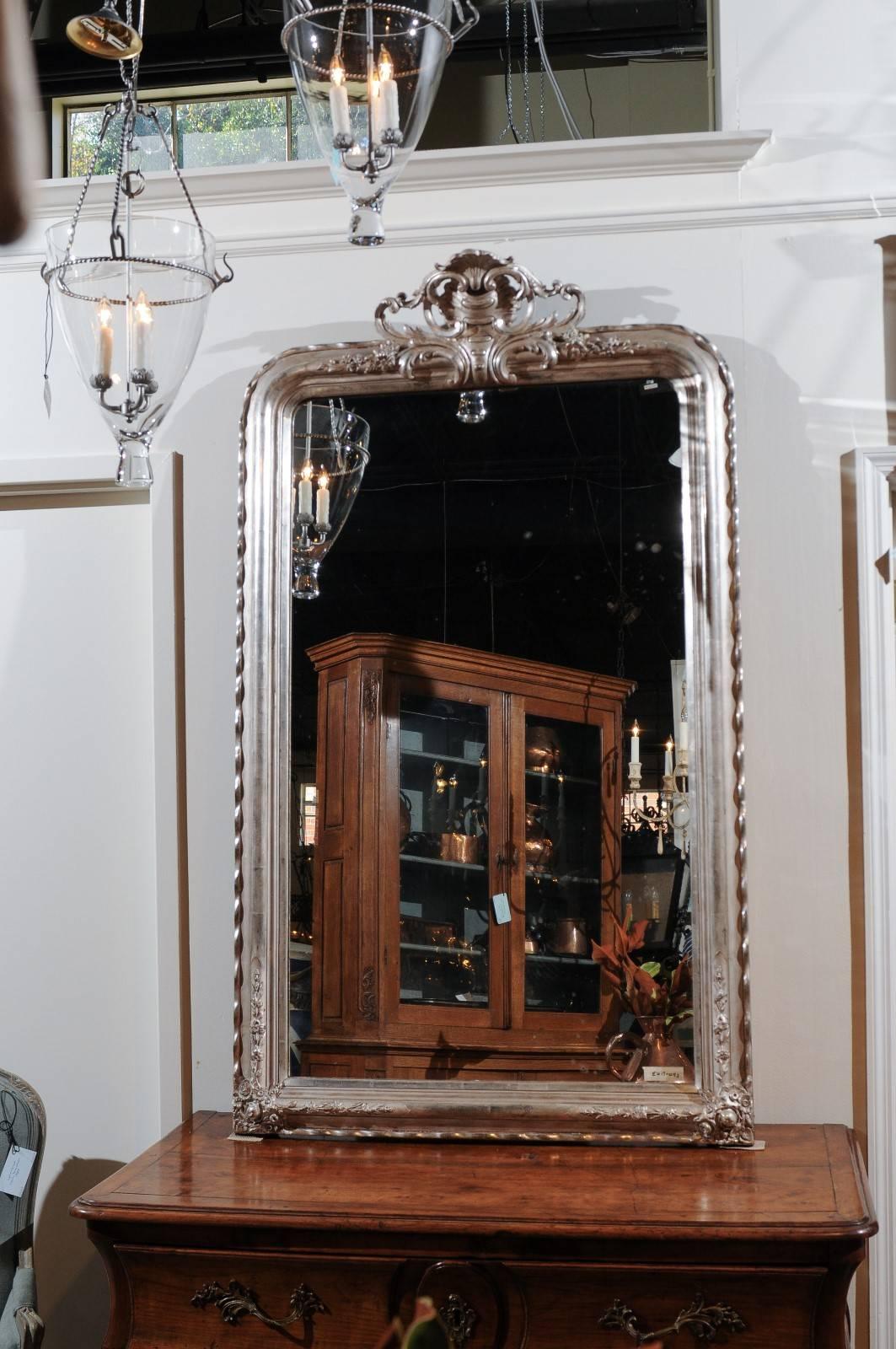 A French Rococo style silver gilt mirror with rocaille carved crest from the late 19th century. This exquisite French mirror features a tall vertical frame, reminiscent in its shape of Louis-Philippe mirrors, beautifully adorned on its crest with an
