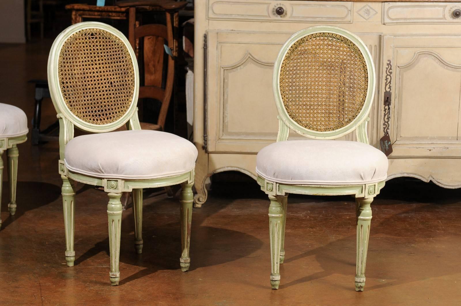 A set of six French Louis XVI style dining room painted chairs from the late 19th century with oval cane backs and new muslin upholstery. Each of this set of Louis XVI style chairs features an oval back with cane upholstery, raised on two delicately