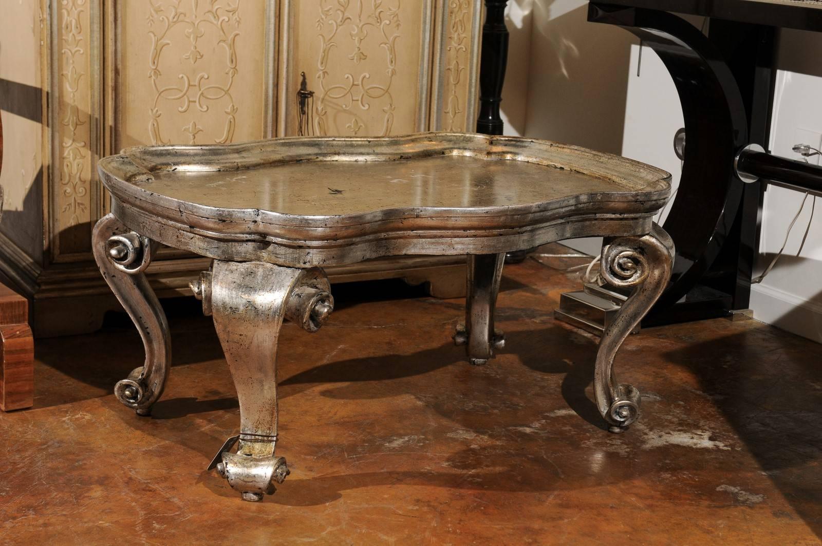An Italian Rococo style silver leaf painted coffee table with tray top from the mid 20th century. This Italian coffee table features a serpentine shaped tray top raised on four exquisitely carved s-scroll legs. The volute motif, particularly praised