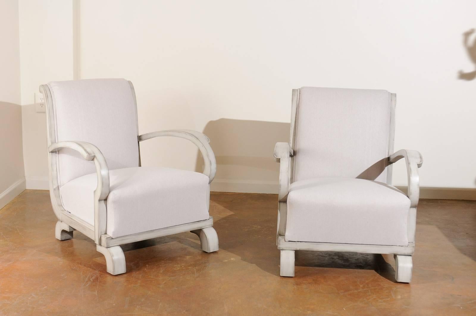 This pair of Art Deco period painted club chairs from the early 20th century features nicely out-scrolled backs and simply curved arms. The chairs are upholstered in a cream fabric. The chairs are raised on four bracket feet turned sideways and