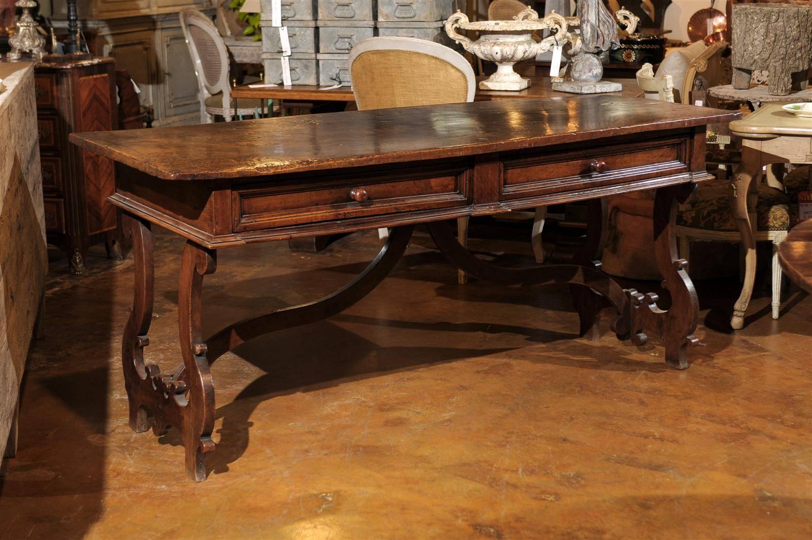 This Italian walnut Baroque style desk from the late 19th century features a rectangular top over two drawers with wooden pulls, resting on a slightly beveled molding. The eye is immediately drawn to the hand-carved lyre shaped legs with their
